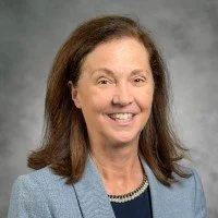 Professional portrait of a woman with shoulder-length brown hair, smiling, wearing a light blue blazer and pearl necklace, against a gray background.