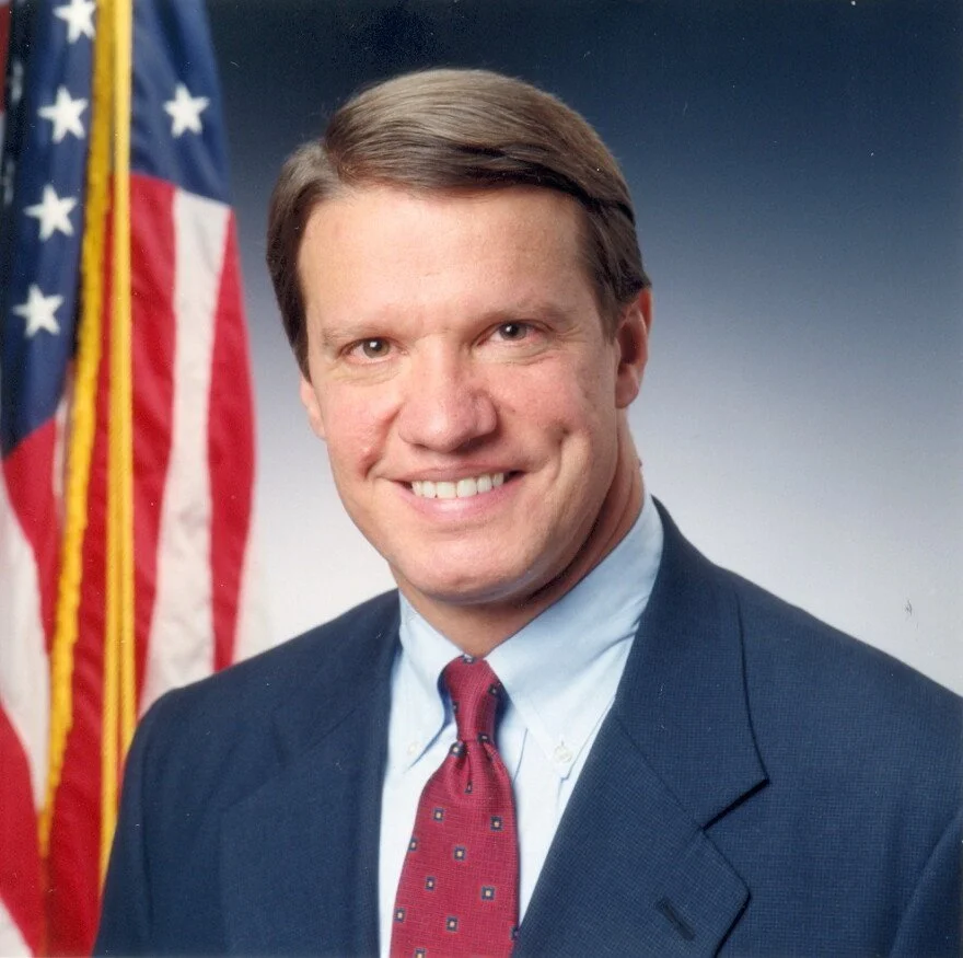 Professional portrait of a man in a suit and tie, standing in front of an American flag.