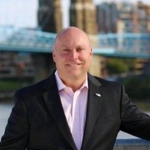 A smiling man with a bald head, wearing a dark suit and light pink dress shirt, standing outdoors in front of a bridge and cityscape.