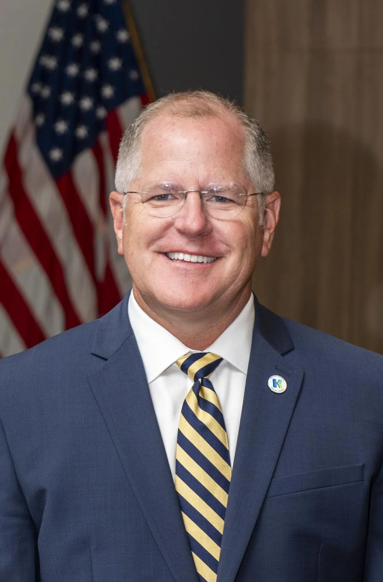 Smiling middle-aged man with glasses in a blue suit and striped yellow and navy tie, standing in front of an American flag.