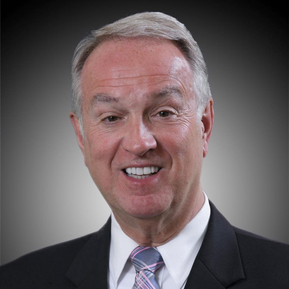 A headshot of a middle-aged man with gray hair, wearing a dark suit, white shirt, and a plaid tie, smiling against a gradient background.