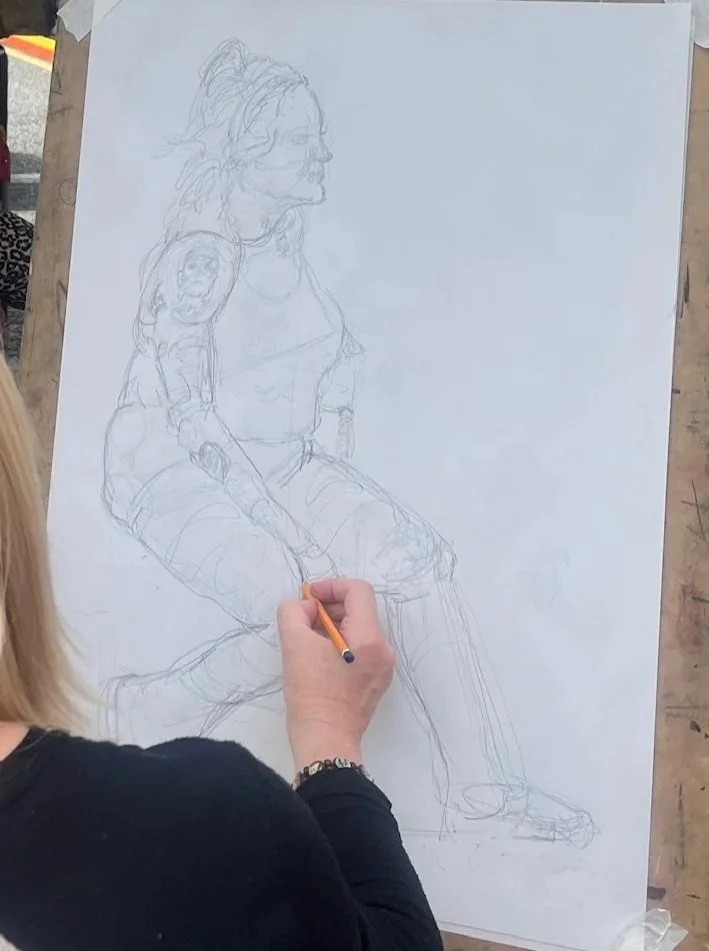 An artist sketching a woman sitting with her arms resting on her lap in a large canvas on an art easel.