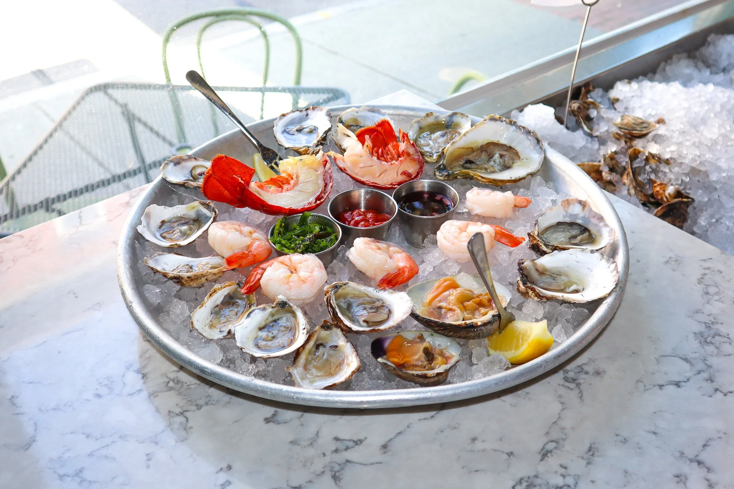 A round metal tray filled with ice, holding oysters on the half shell, cooked shrimp, lemon wedges, and small containers of cocktail sauce and soy sauce, with green onions and hot sauce in the center.