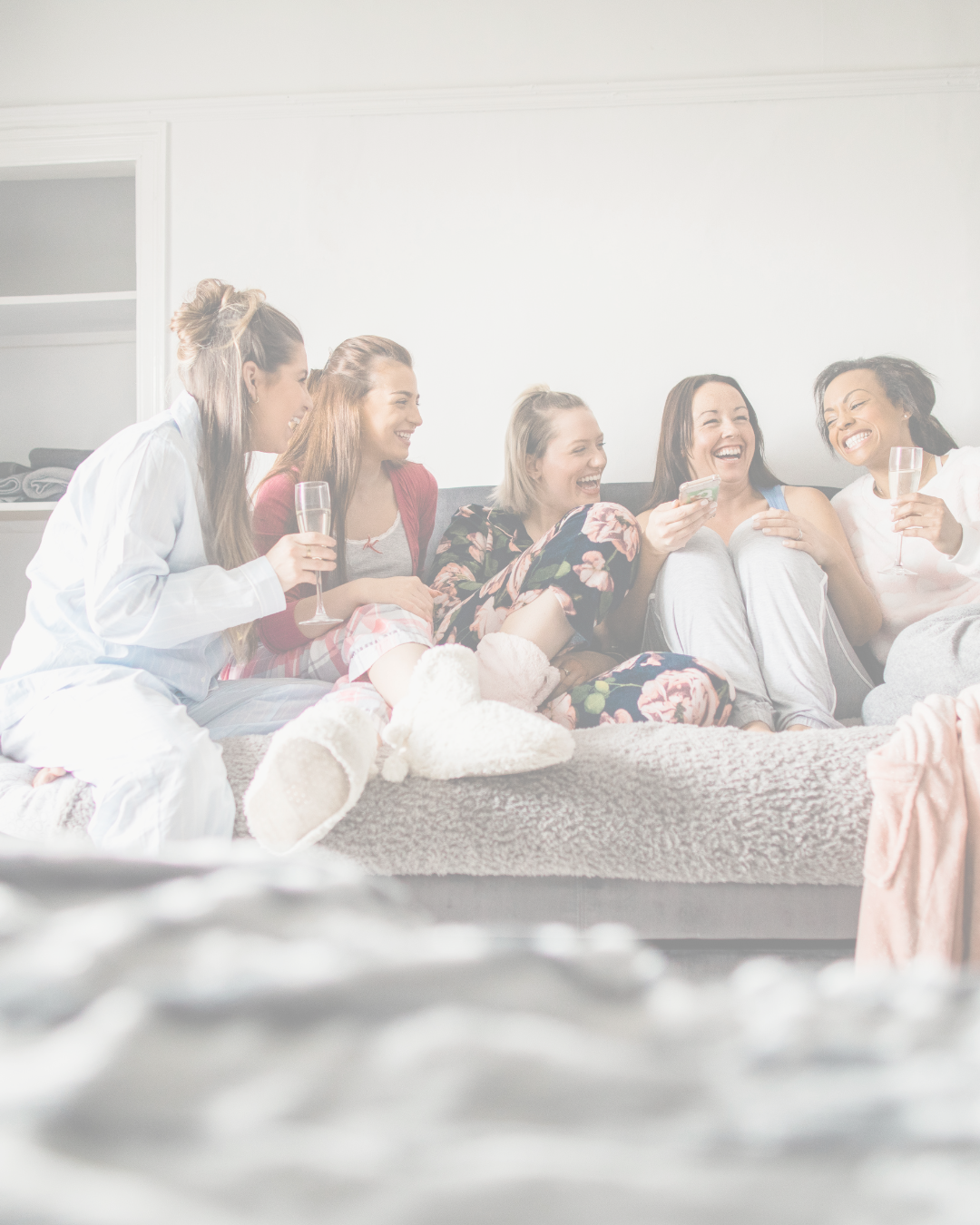 A group of six women sitting on a couch, laughing and enjoying each other's company, some holding glasses of champagne, in a bright room.