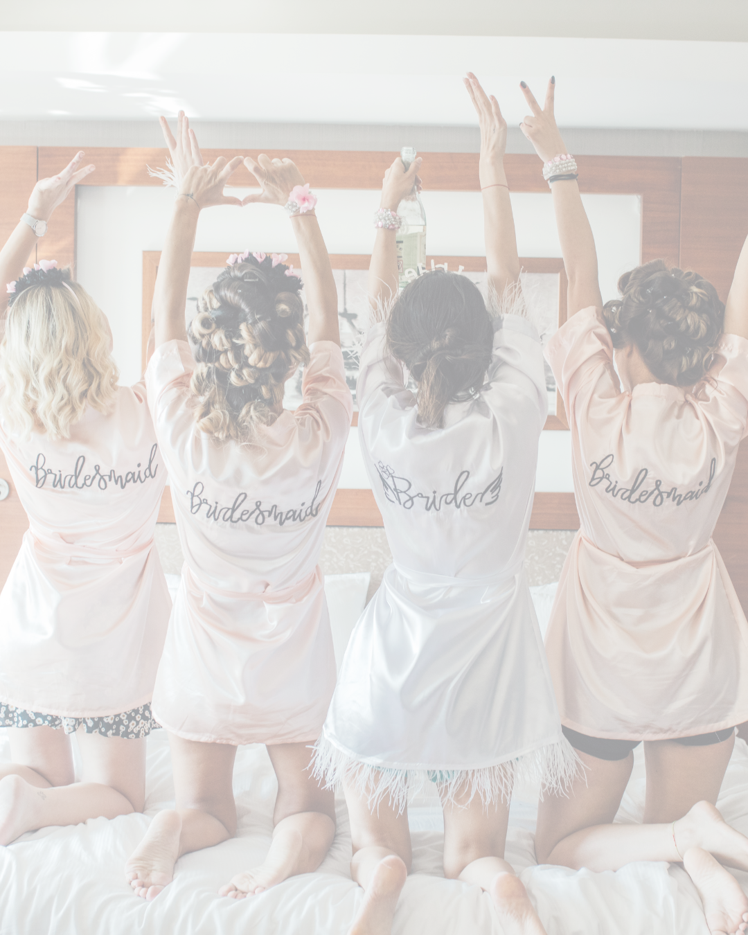 Four women in satin robes with 'Bridesmaid' written on the back, kneeling on a bed with hair rollers, celebrating with raised hands, in a hotel room before a wedding.