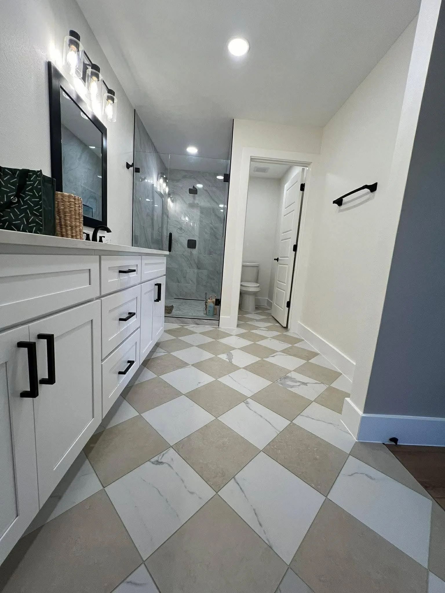 Modern bathroom with white vanity, black hardware, large mirror, glass-enclosed shower, checkerboard tile floor, and toilet room