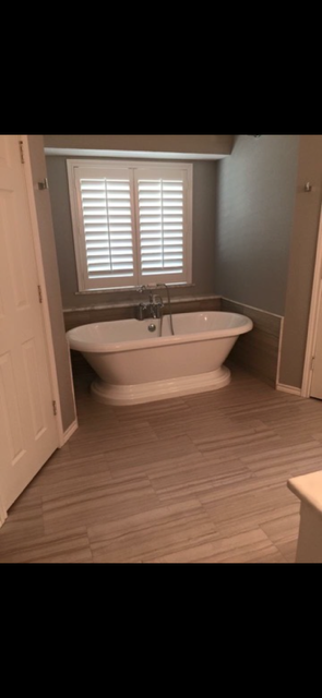 Freestanding white bathtub beneath plantation shutters in a modern bathroom with wood-look flooring