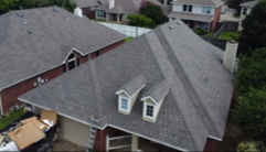 Aerial view of a gray shingle-roofed suburban house with dormer windows and neighboring homes