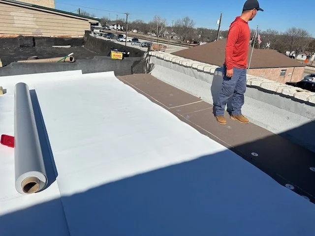 Man in a red shirt standing on a rooftop beside a large roll of white roofing membrane