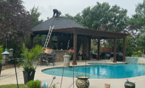 Landscaped backyard with a turquoise swimming pool, covered pavilion, potted palms, and a person on a ladder repairing the roof