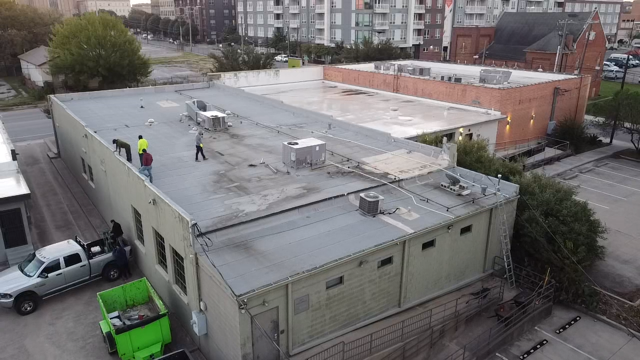 Aerial view of workers maintaining a flat commercial rooftop with HVAC units, a green dumpster, and surrounding apartment buildings.