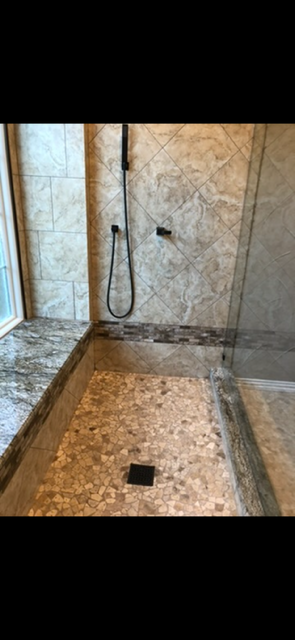 Modern walk-in shower with beige stone tile walls, handheld showerhead, mosaic tile floor, and black square drain.