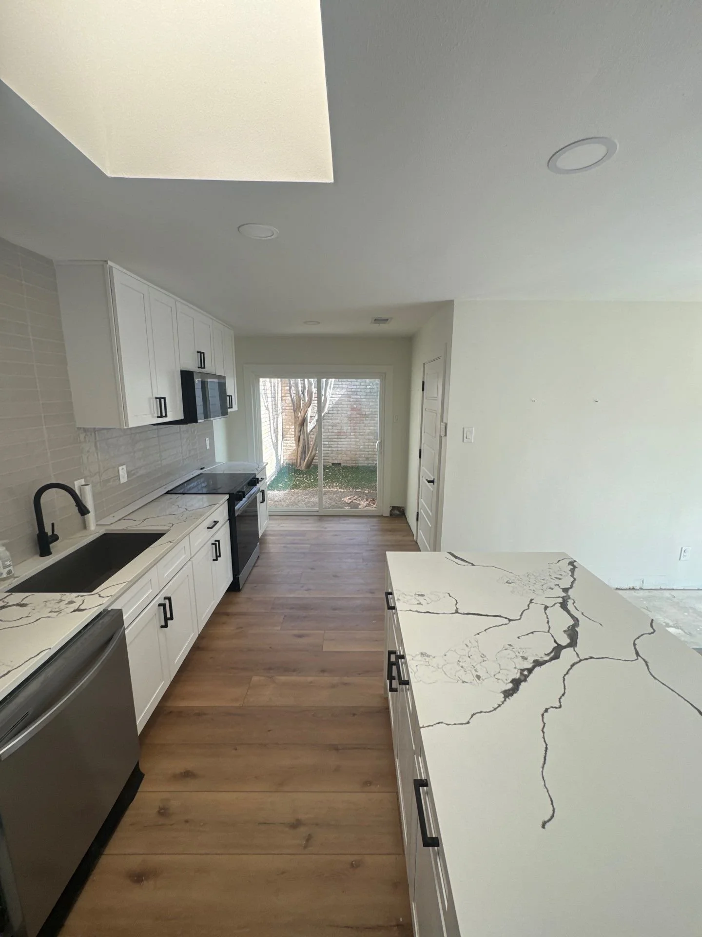 Modern white kitchen with marble-look countertops, black fixtures, stainless steel appliances, wood flooring, and sliding glass doors to a backyard