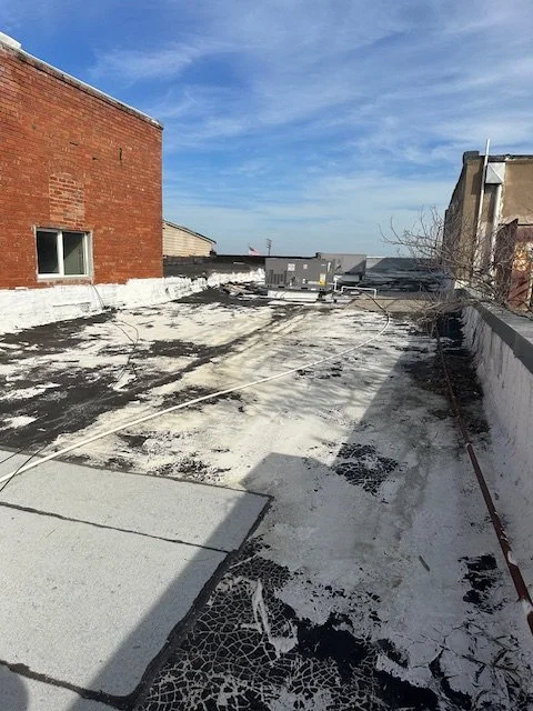 Flat rooftop with peeling white coating, exposed black roofing, utility equipment, and brick buildings under a blue sky