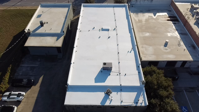 Aerial view of three flat-roofed commercial buildings with white roofs, HVAC equipment, parking areas, and surrounding streets.