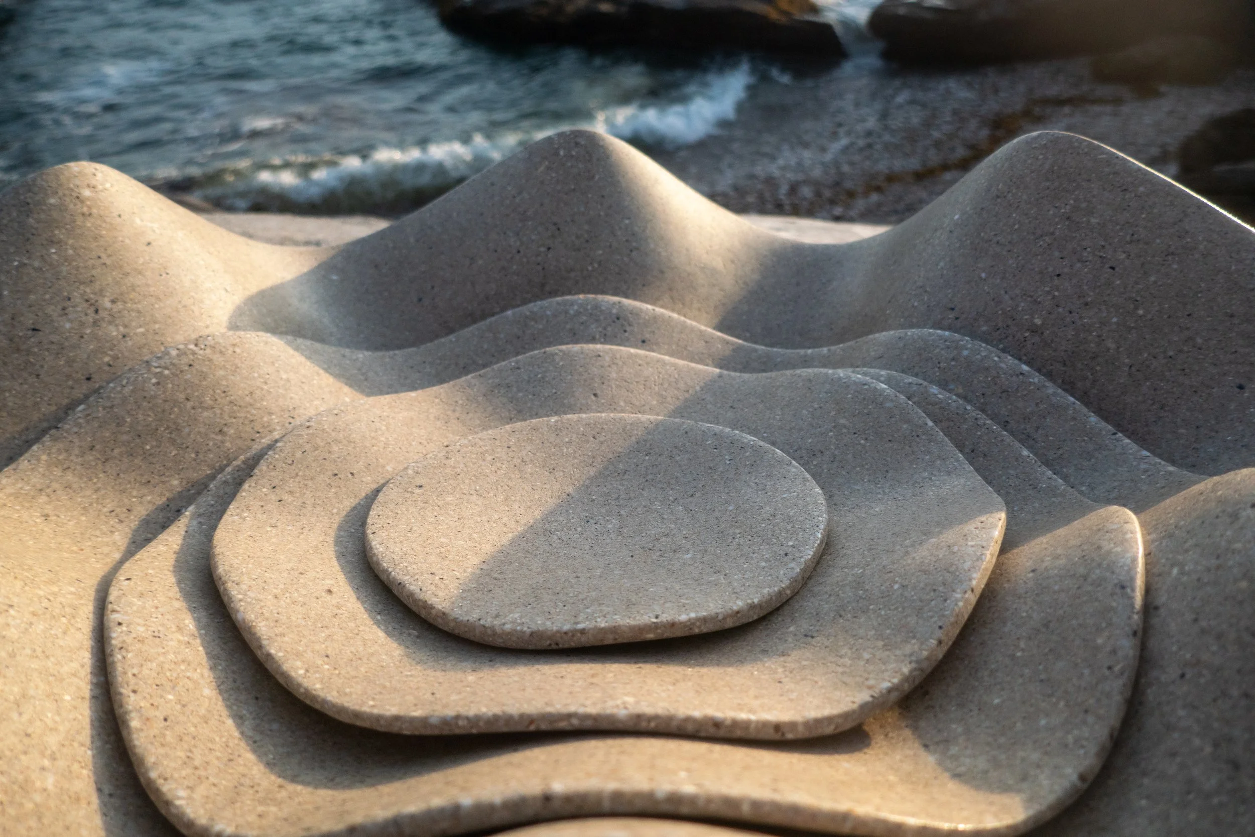 Close-up of Clam Family of Ripple bowls arranged in a curved line on a sandy beach, with the ocean and gentle waves in the background.