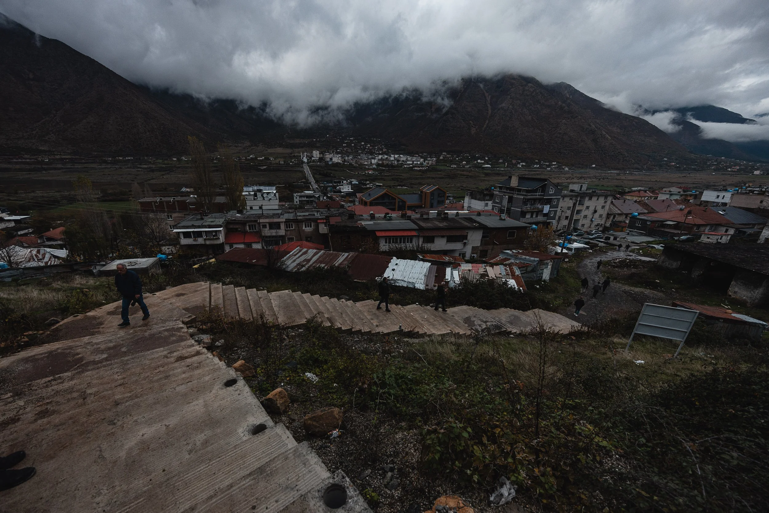People walk up the stairs heading to the mine, with the city visible in the background in Bulqizë, Dibër County, Albania on November 11, 2025. The image captures miners commuting to work in one of Europe’s largest chromium mining areas, central to th