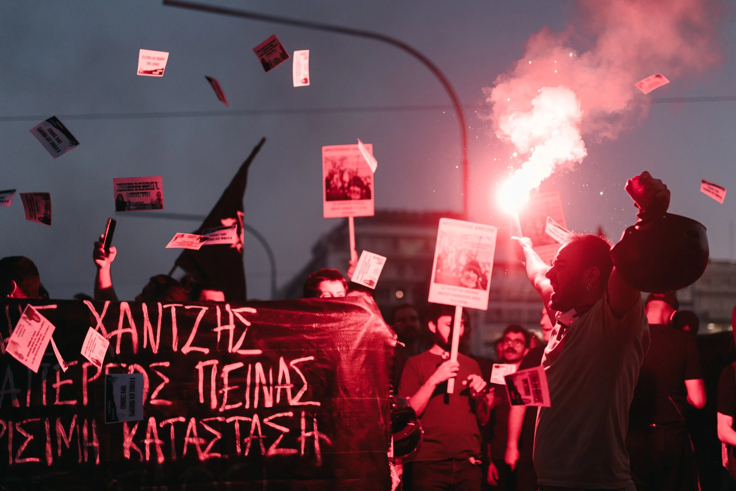 A man holds a red flare in front of protesters holding a large banner as flyers fill the air during a protest in Athens, Greece on June 23, 2026. The protest was held in support of the Prosfygika community, a historic refugee housing complex and symb