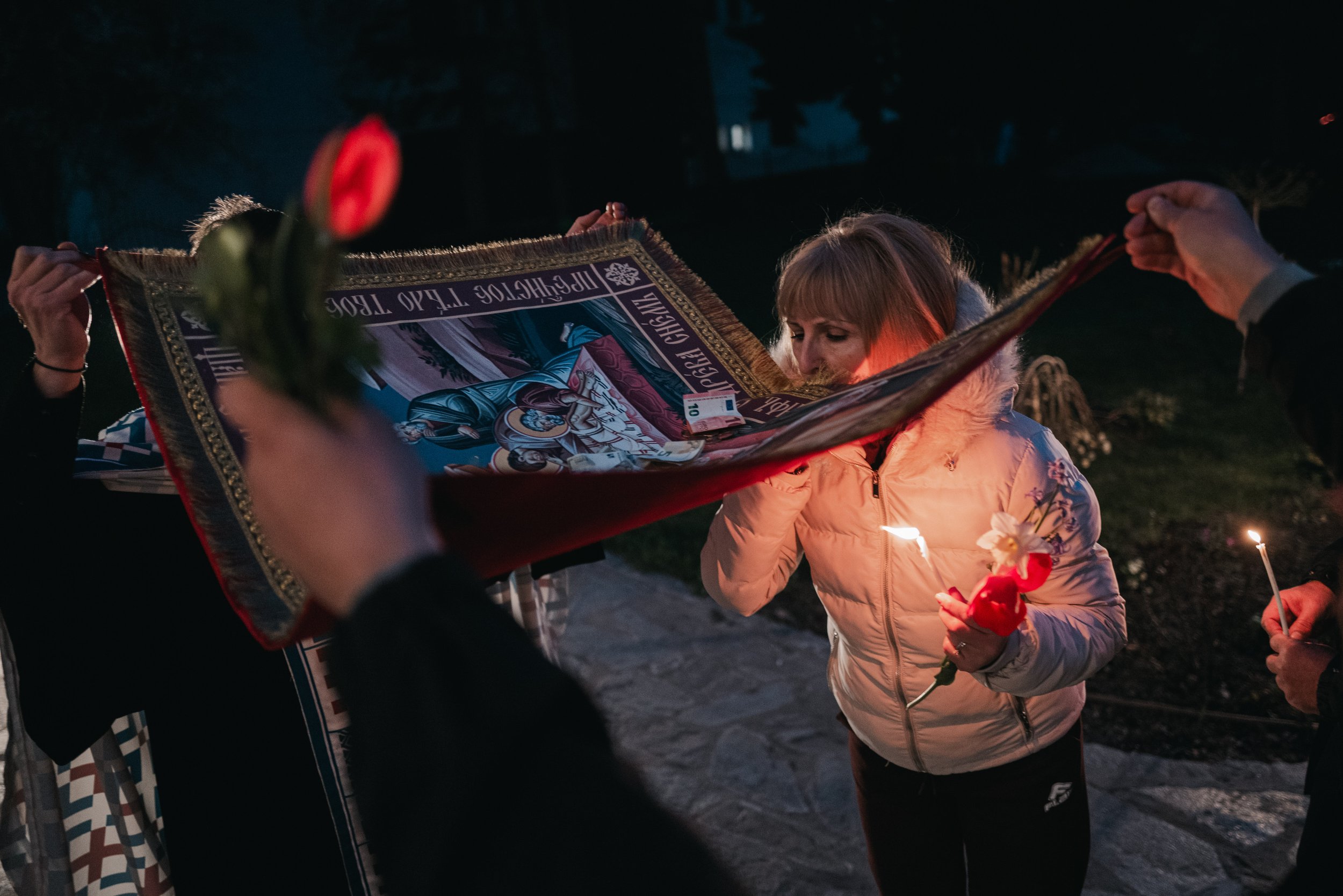 A woman holding flowers and a candle kisses the Holy Shroud raised by a priest and his assistant before passing beneath it in Iskrets, Bulgaria on April 10, 2026. The ritual is part of the Orthodox Good Friday service, symbolizing the burial of Jesus