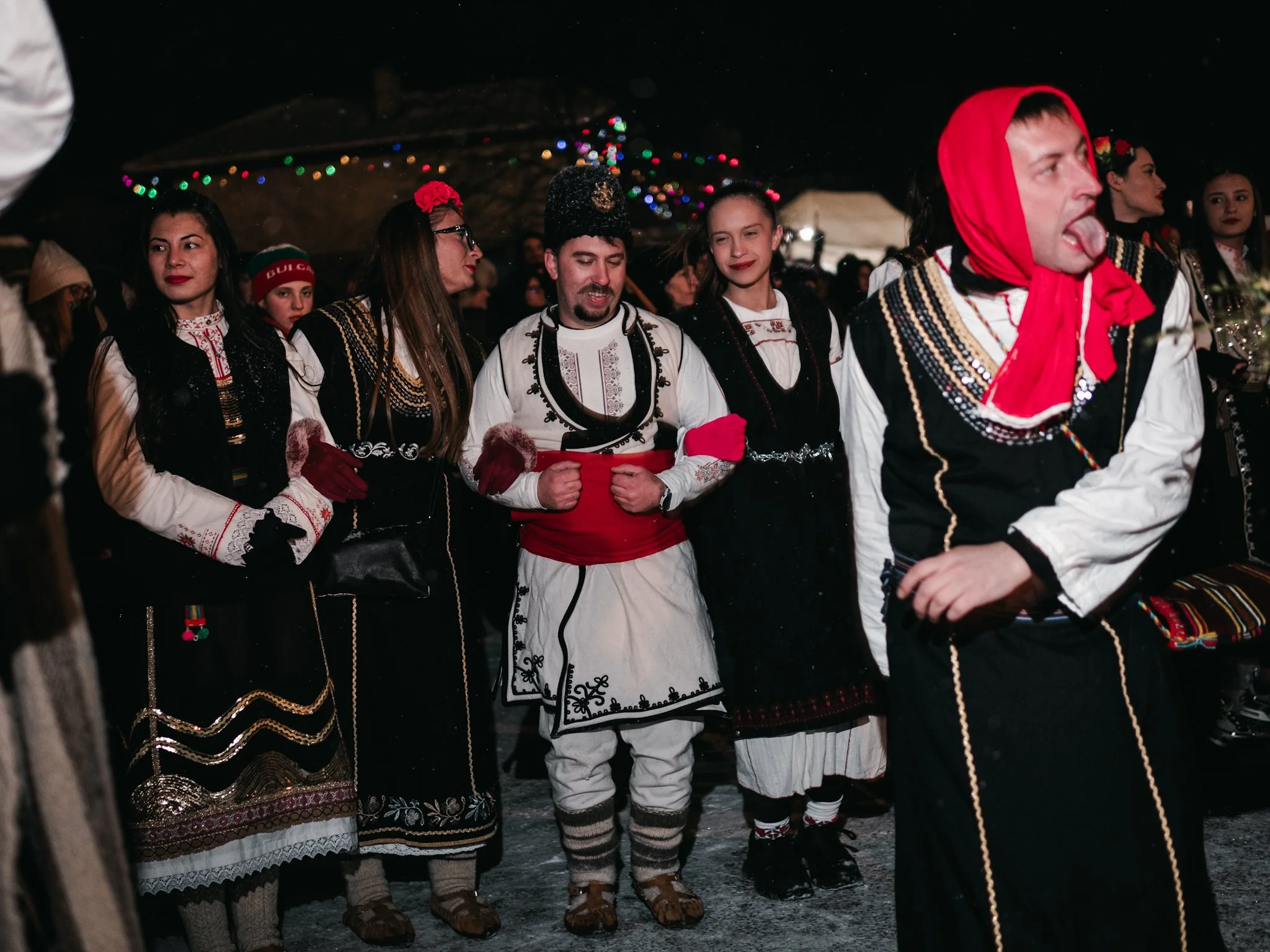 In the early Surva traditions, roles such as the woman’s character were also performed by men. Here, a man dressed in traditional women's clothing, playfully sticking out his tongue, stands before a group of three women and another man, embodying the