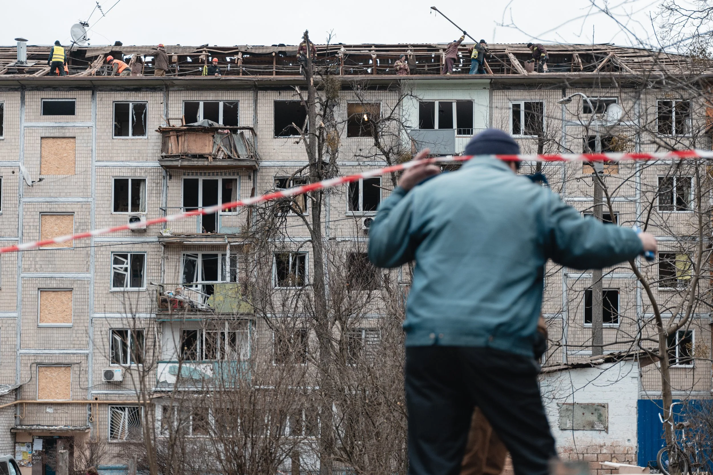 A man lifts the red and white tape cordoning off the struck residential building while others clean the roof in the Sviatoshynskyi district of Kyiv, Ukraine on December 23, 2025. The incident occurred during a Russian aerial strike that partially dam