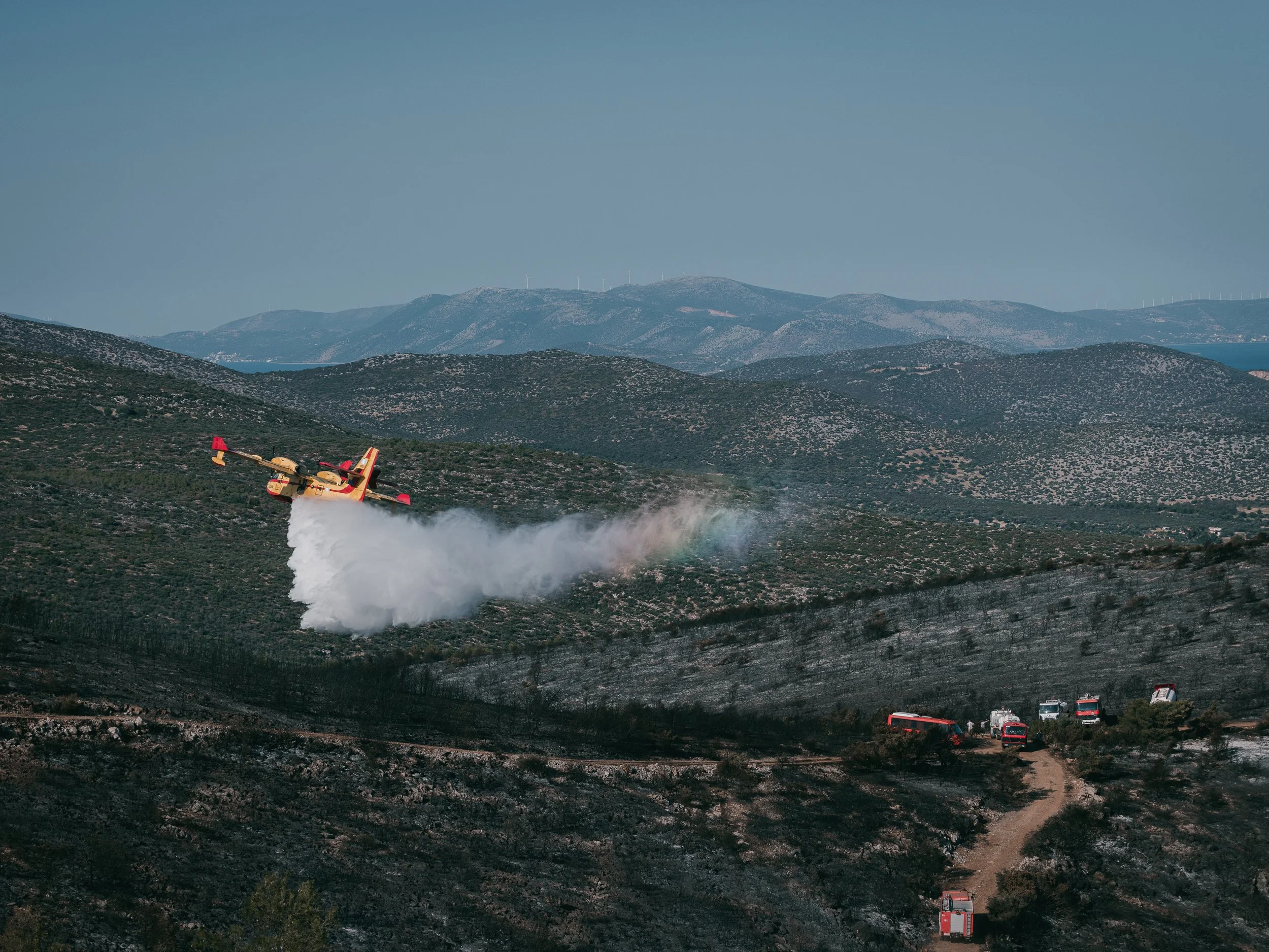 A firefighting plane releases water over a forest as white firefighting trucks stand by on the ground in Ano Souli, near Athens, Greece, on June 16, 2025