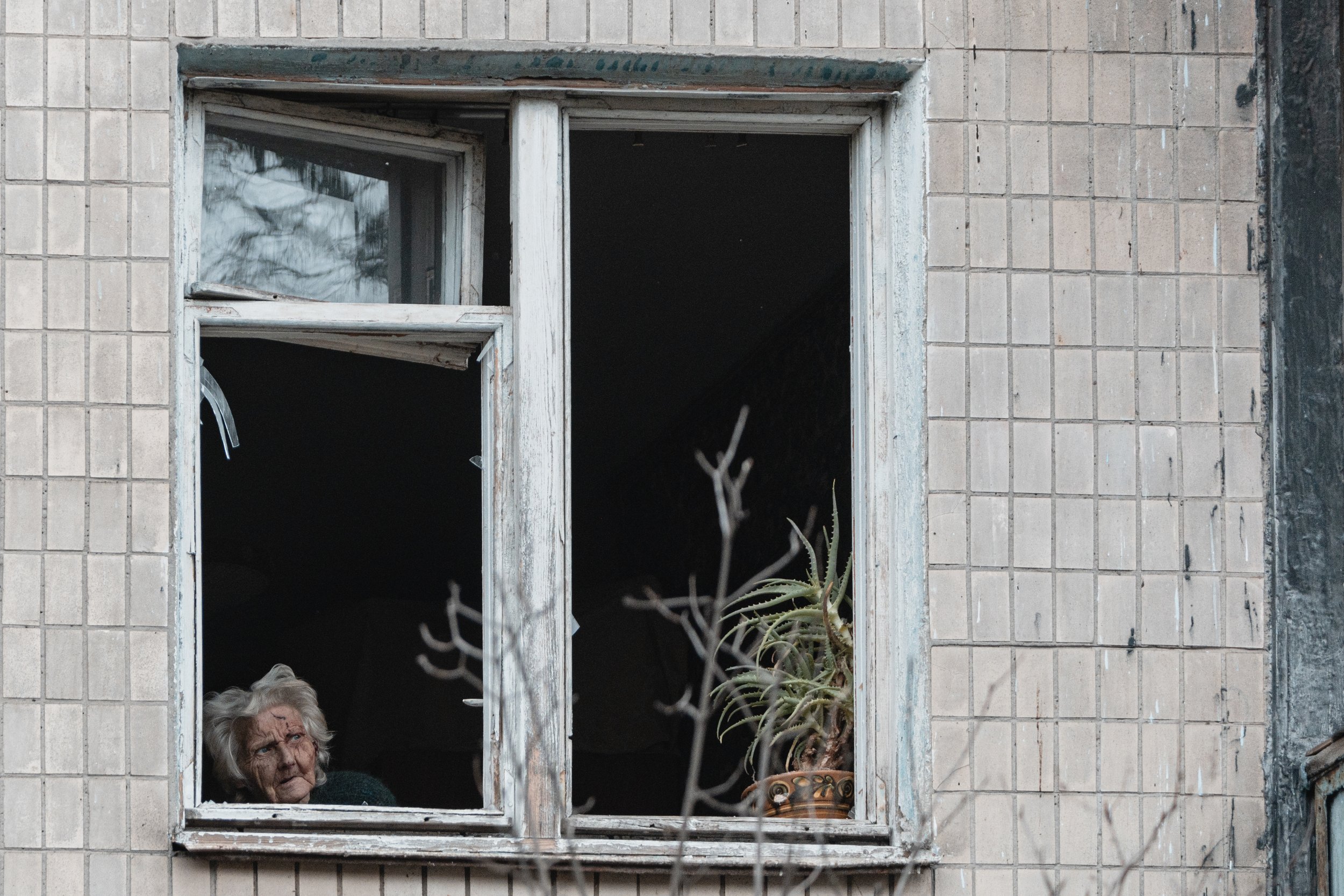 An elderly woman looks through her broken window towards the damaged residential building in the Sviatoshynskyi district of Kyiv, Ukraine on December 23, 2025. The incident occurred during a Russian aerial strike that partially damaged the building’s