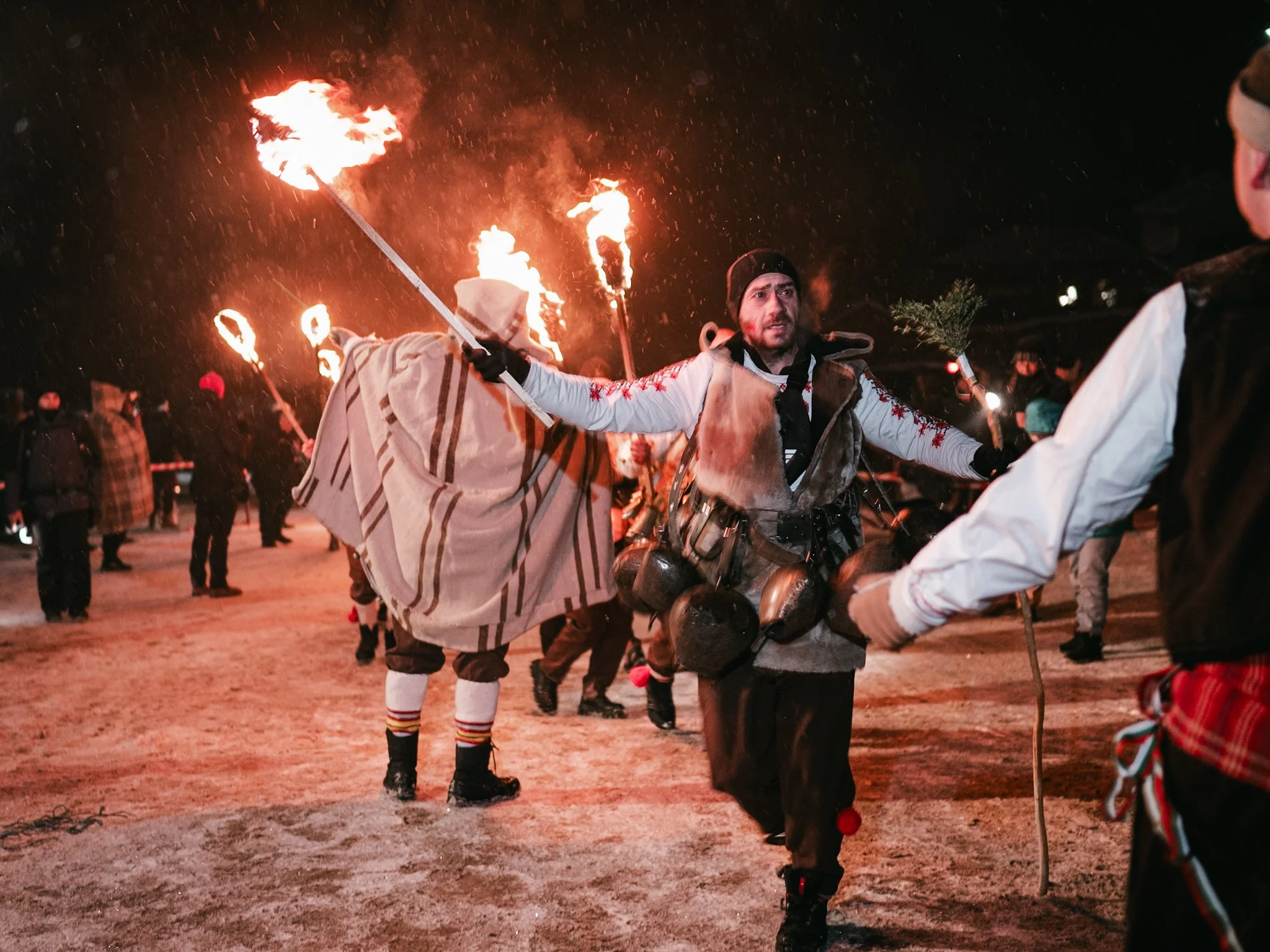 A man, adorned with traditional Surva bells, performs a lively dance while holding a torch, embodying the spirit of the Kosharevo festival and its rituals to ward off evil and bring good fortune.