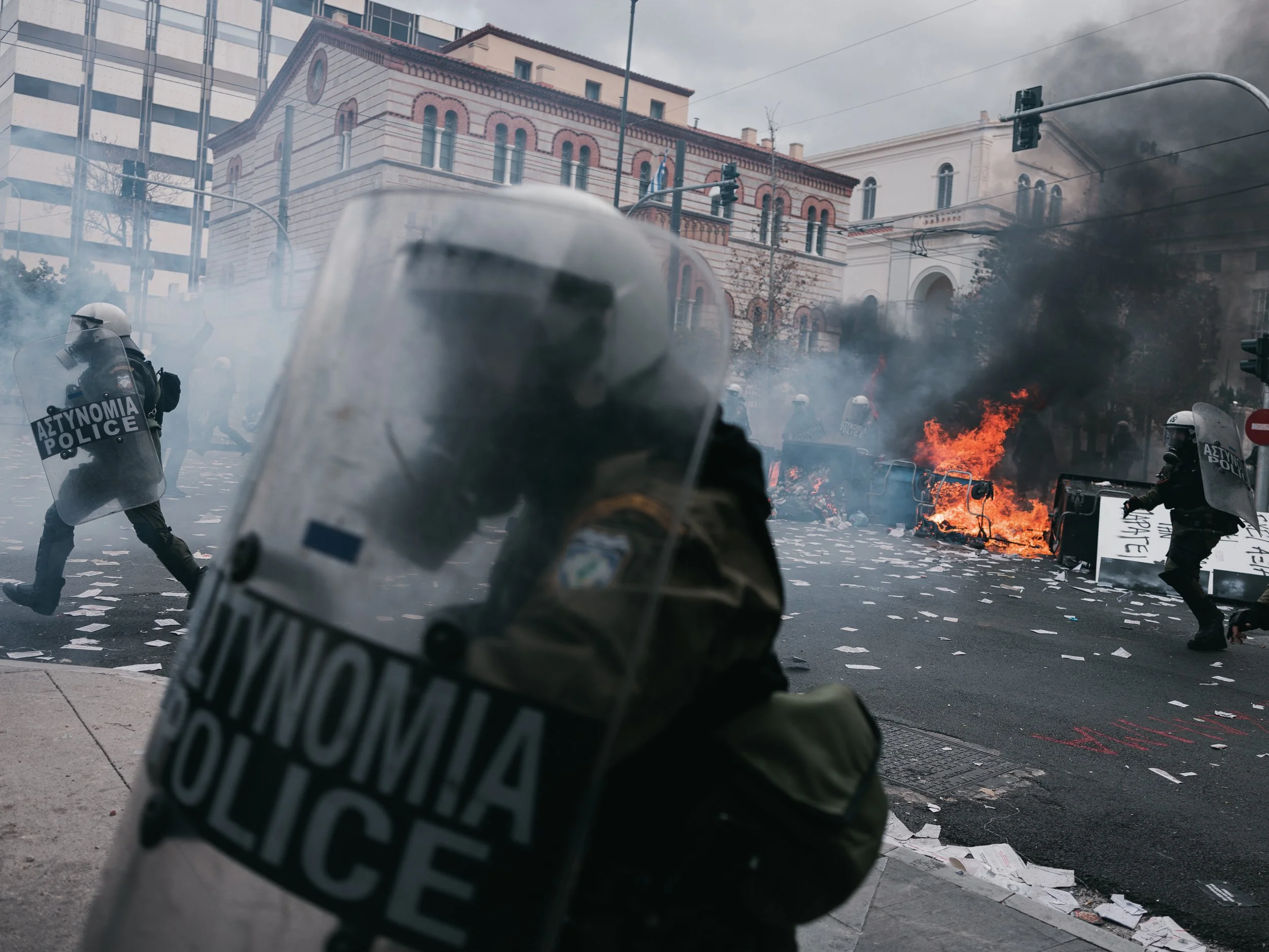 Protesters create a barricade with a burning trash bins on a crossroad as police officers run toward them and throw tear gas, forming a line in the picture, in Athens, Greece on February 28th, 2025.