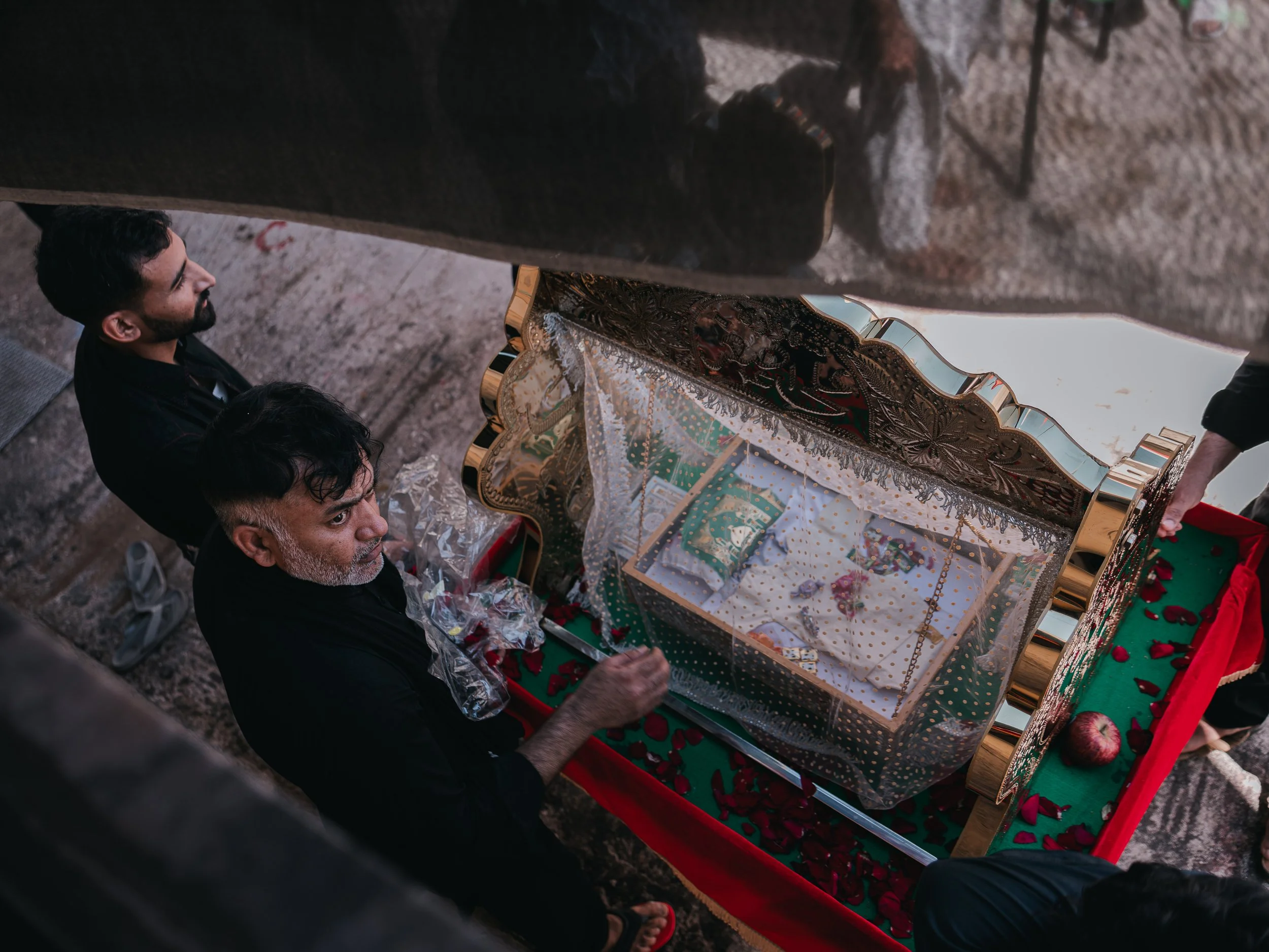 An overhead view shows a child’s coffin, symbolizing Ali Asghar, the youngest son of Imam Husayn who was martyred at Karbala, with men standing nearby during Ashura preparations. July 2025, Peristeri, Athens, Greece.