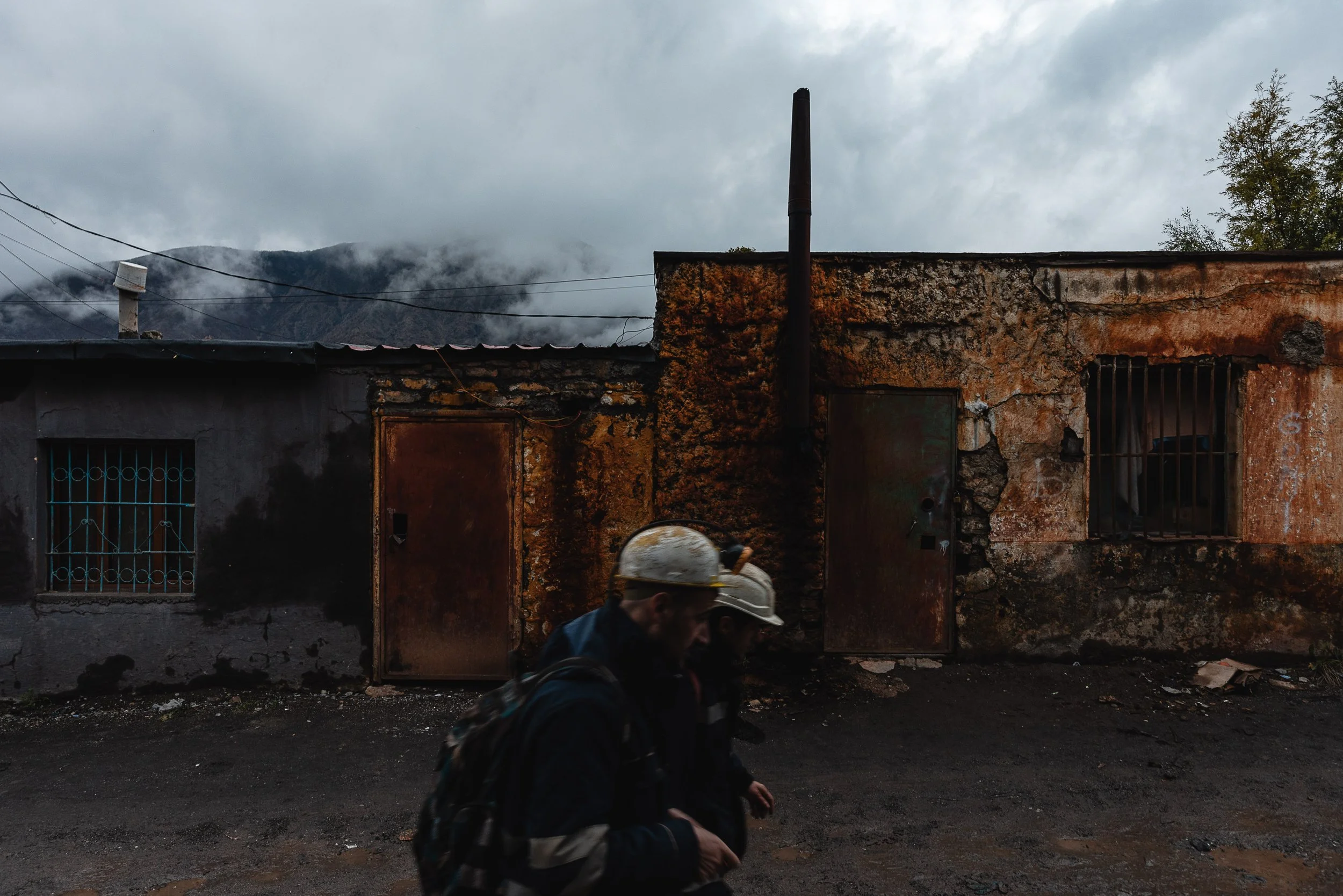 Men return from work in the mine, walking in front of old buildings in the industrial complex in Bulqizë, Dibër County, Albania on November 11, 2025. The scene highlights the daily life of miners in a town shaped by decades of chromium extraction.