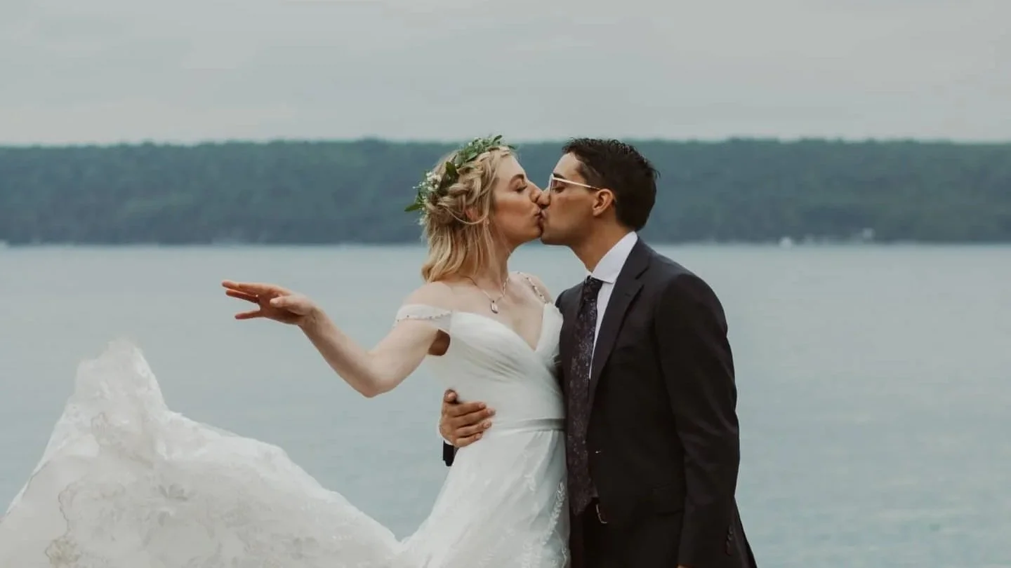 A bride and groom kissing by a lake. The bride is in a white wedding dress and flower crown, and the groom in a black suit, holding her by the small of her waist.