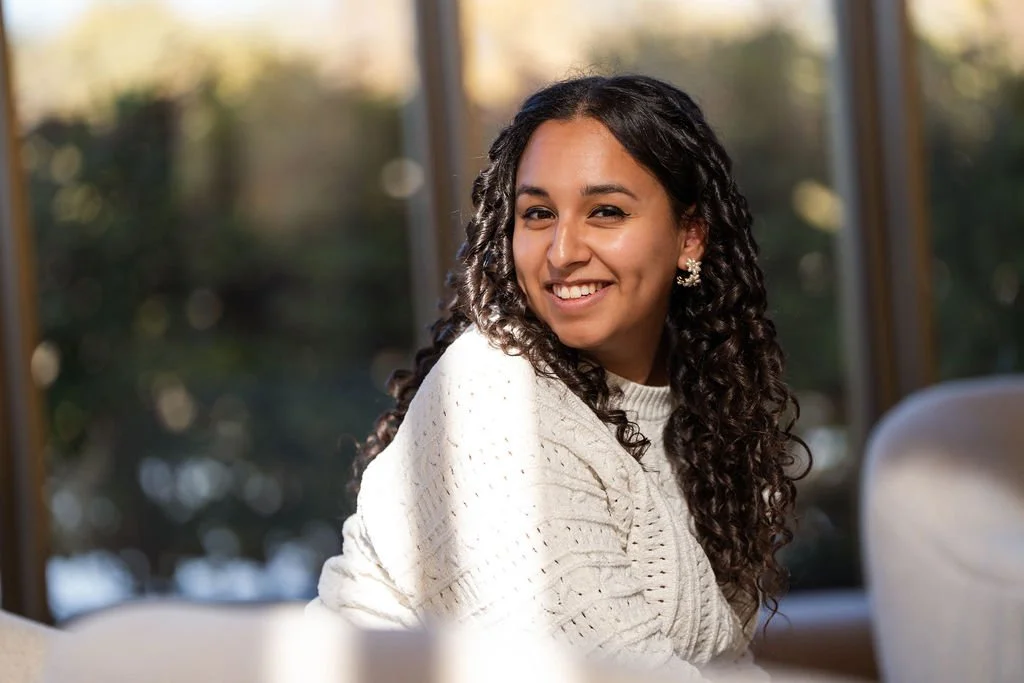 Young woman with long curly hair smiling in a cozy indoor setting with natural light.