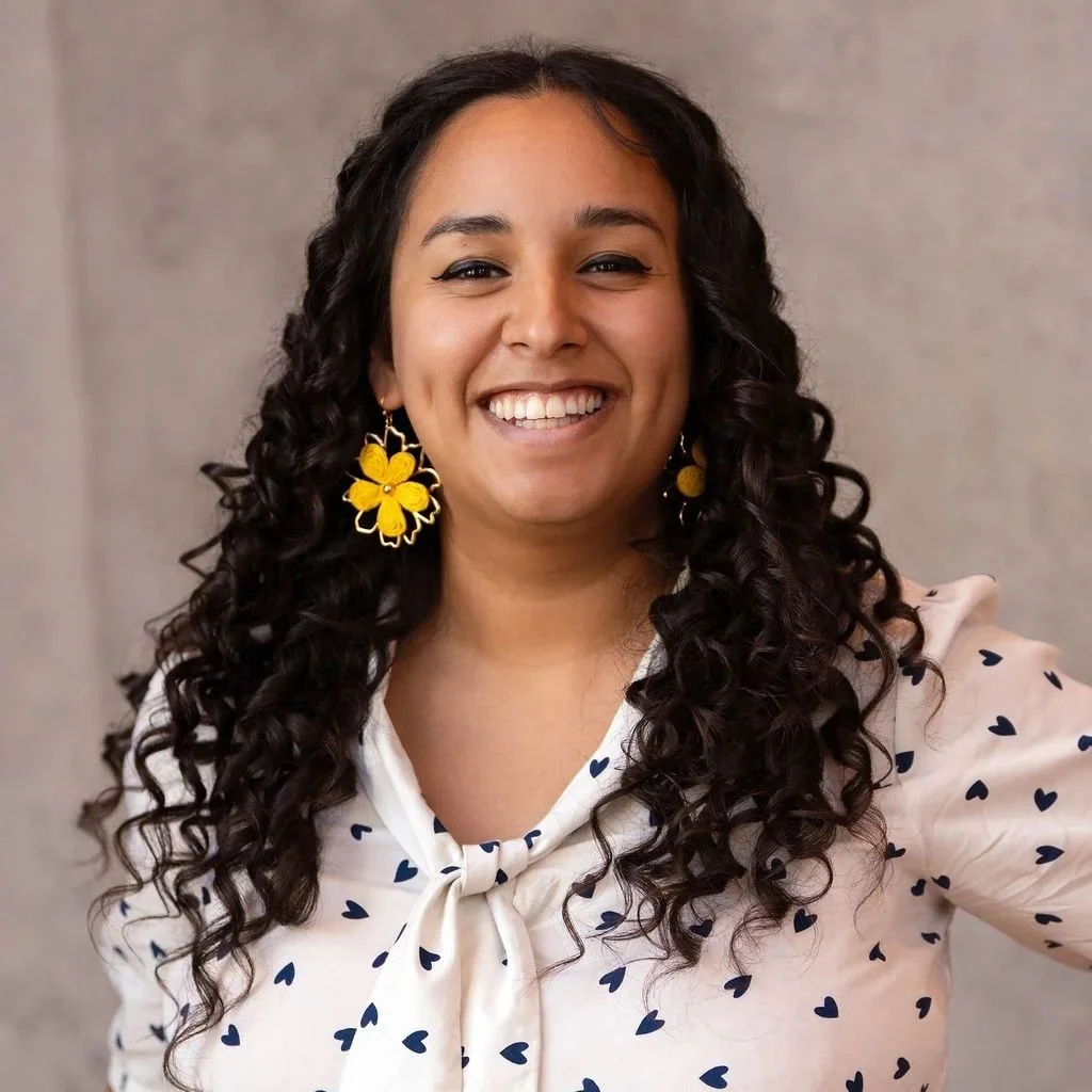 A woman with curly dark hair, wearing yellow floral earrings, a white blouse with dark blue hearts, and smiling at the camera.