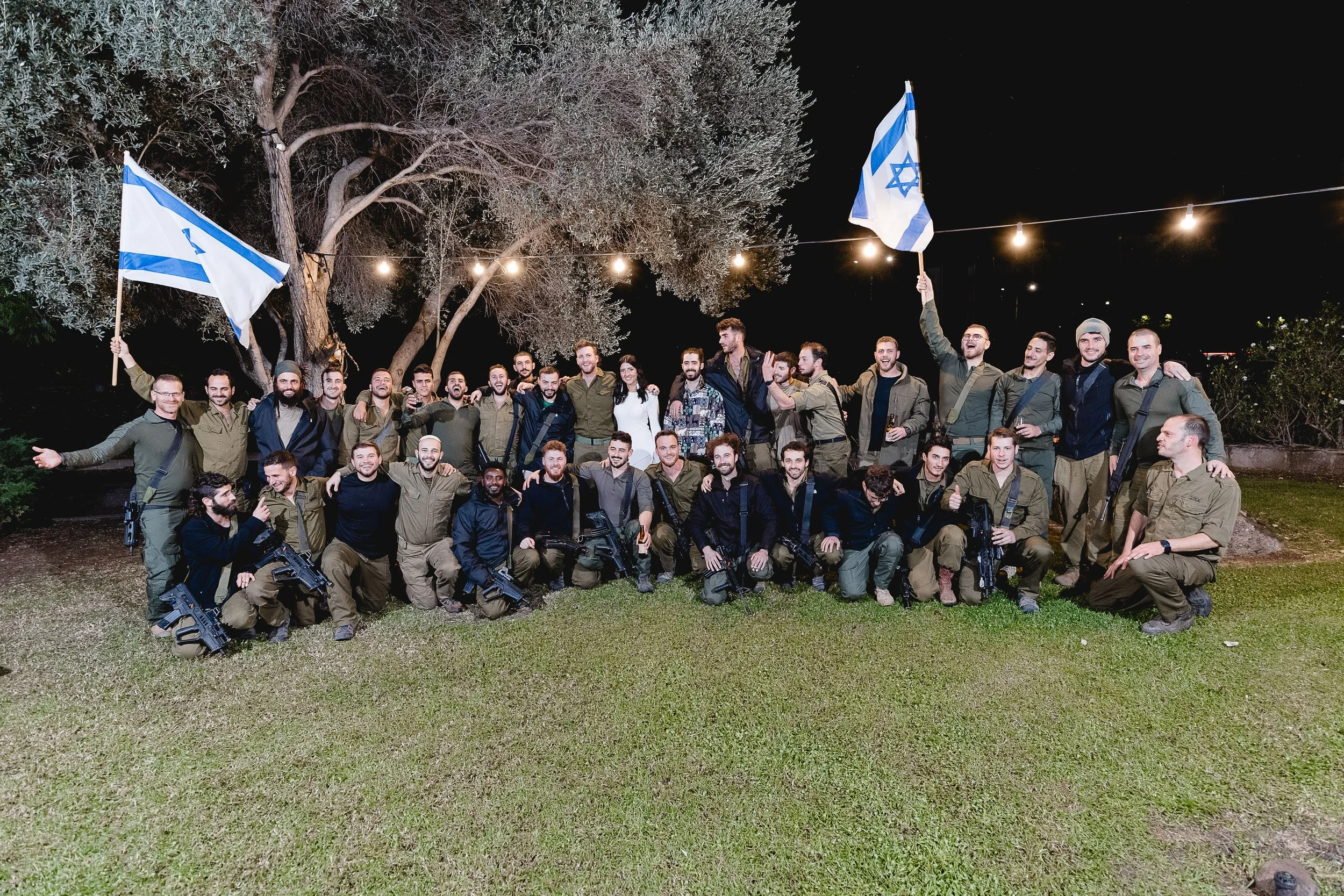 Group of soldiers posing outdoors at night, some holding Israeli flags, with string lights overhead.