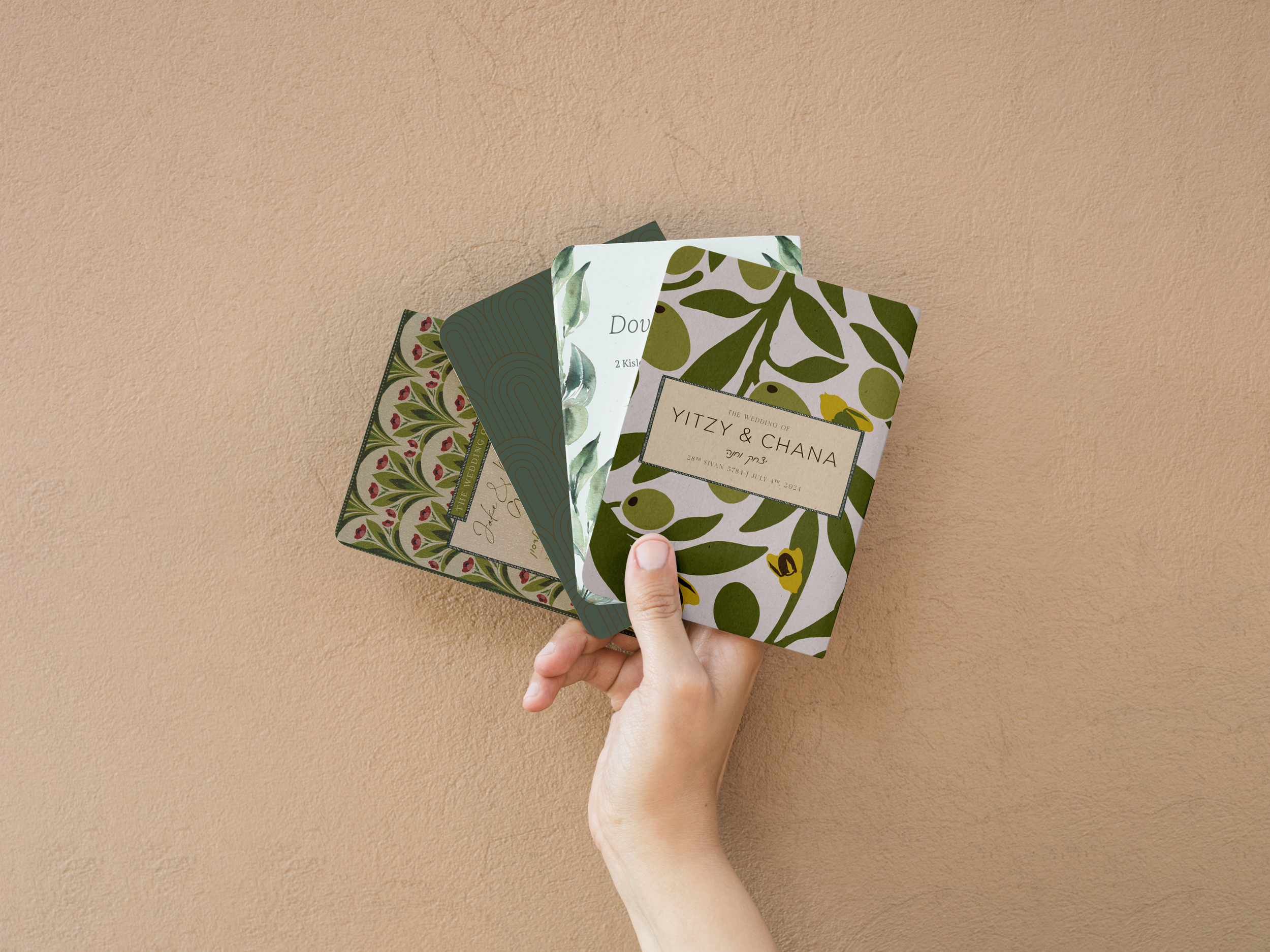 Four intricately designed wedding invitations with floral and botanical patterns being held in a person's hand against a beige textured background.