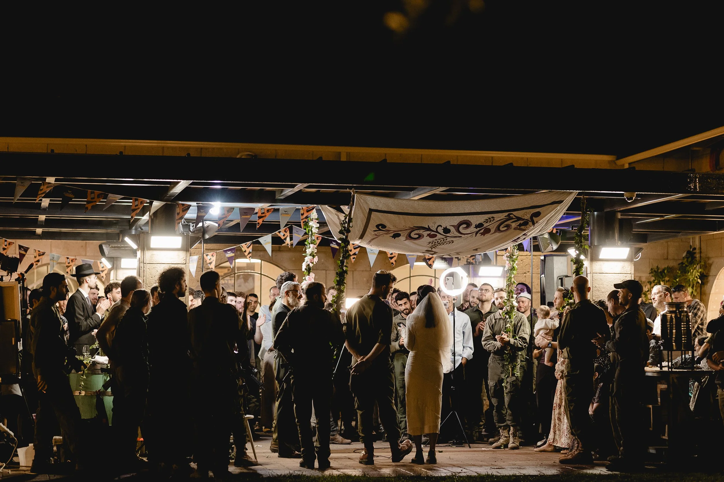 Nighttime wedding reception with guests gathered outdoors, bride and groom in center, string lights, bunting decorations, and festive ambiance.