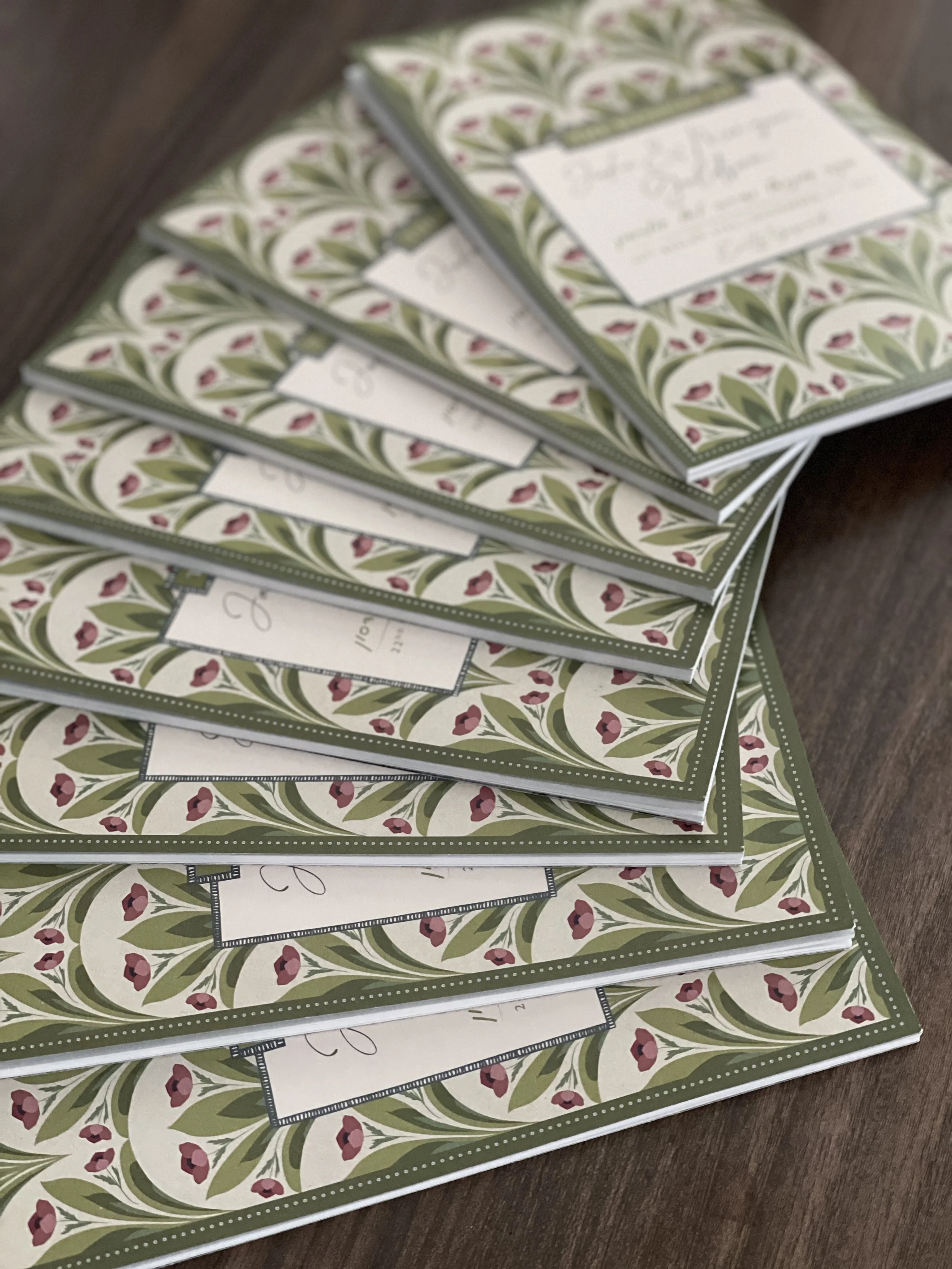 Stacked invitation cards with green, white, and red floral design on a dark wooden surface.