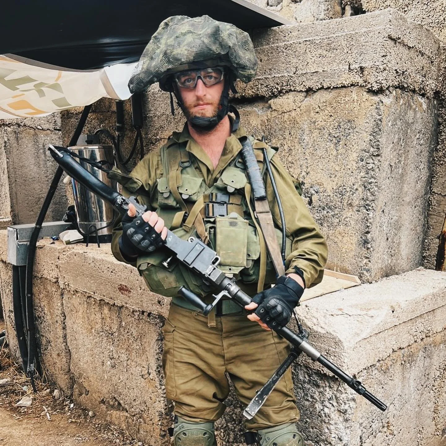 A soldier in camouflage uniform with tactical gear holding a machine gun, standing against a concrete structure.