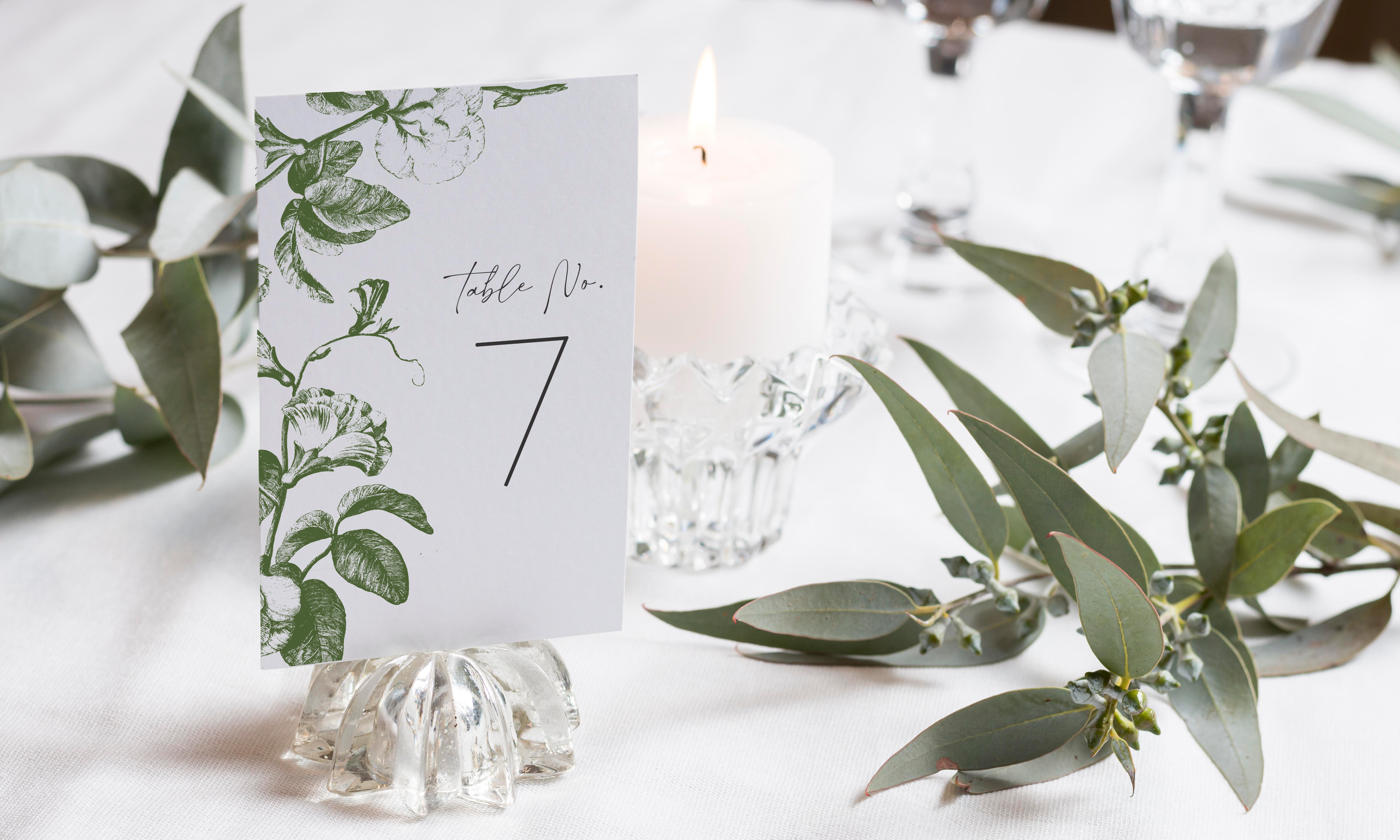 Wedding reception table setting with a table number card labeled 7, a white candle in a decorative glass holder, greenery with leaves, and water glasses on a white tablecloth.