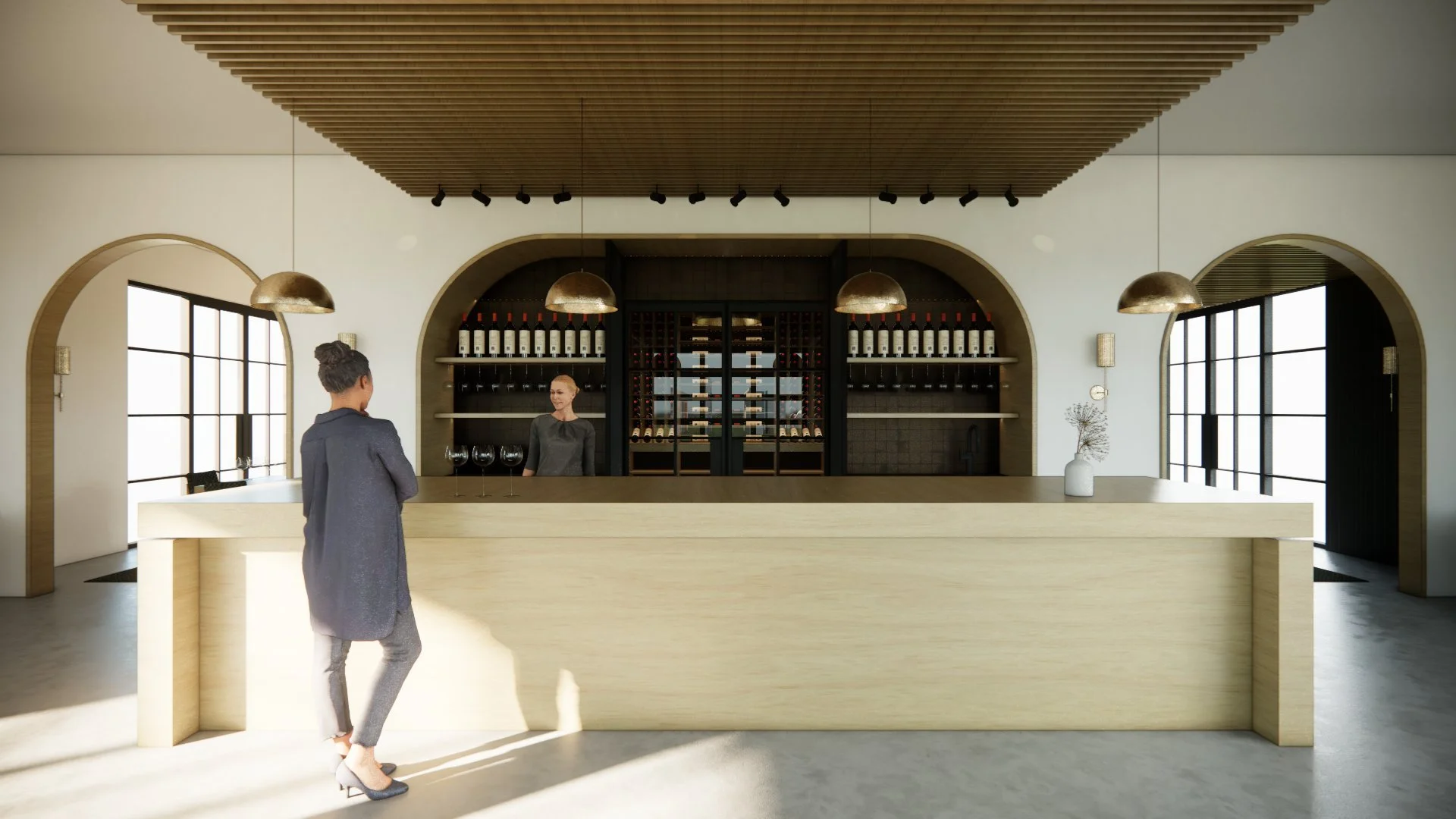 Interior of a modern wine tasting room with a wooden counter, bottles of wine on shelves behind, and large windows on either side. Two women are present, one standing behind the counter and the other in front, engaging in conversation.