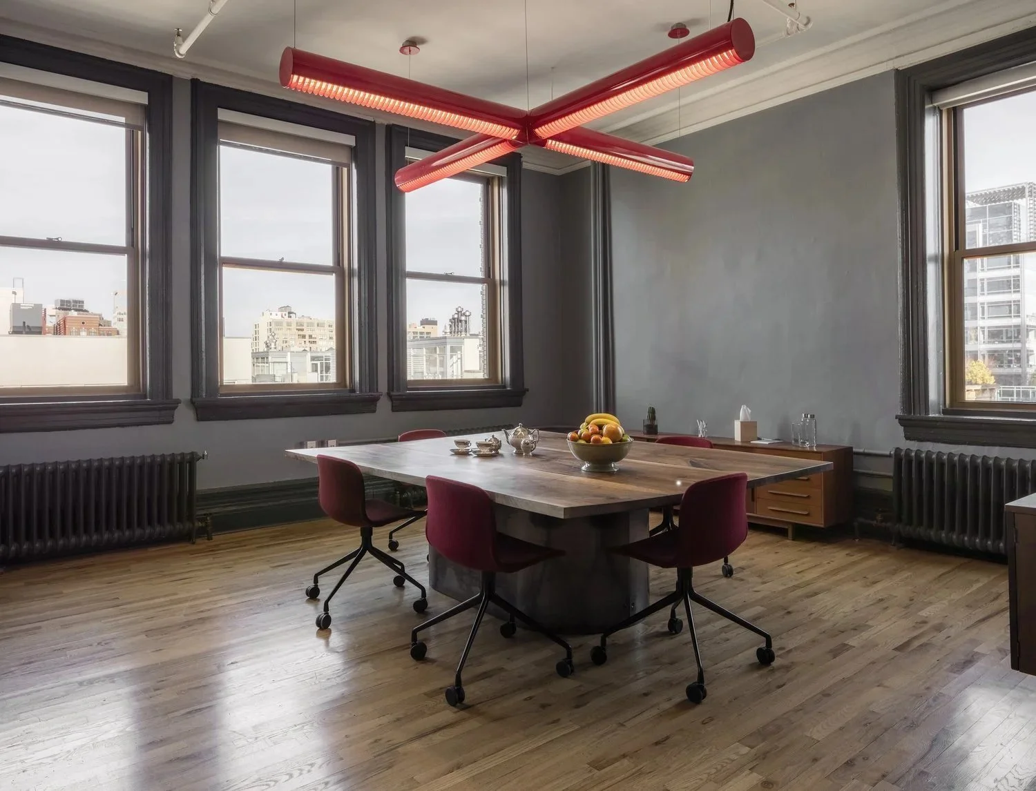 Empty modern conference room with a large wooden table, red chairs, multiple windows with city views, wooden floors, and a unique red ceiling light fixture.