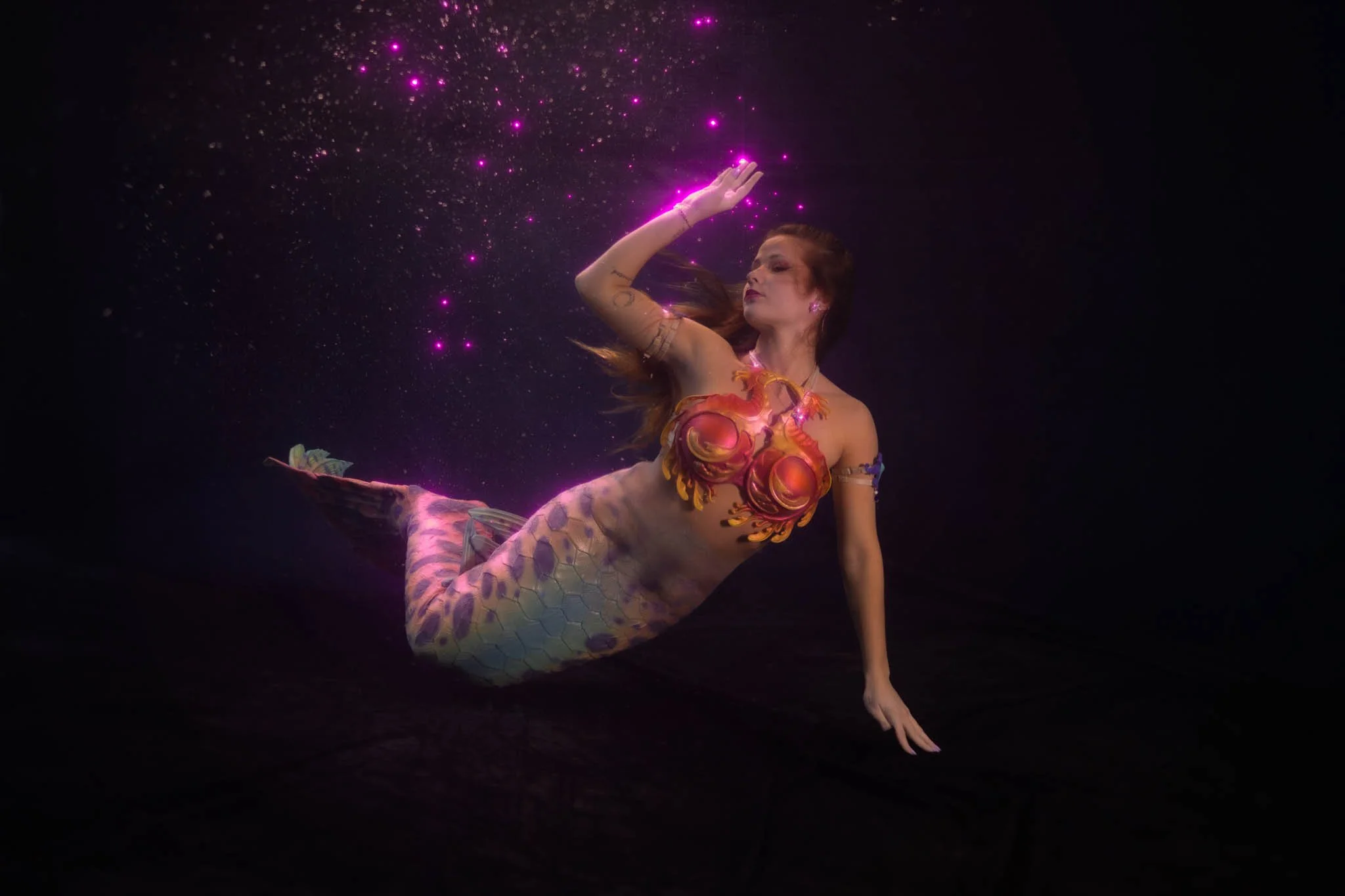 A woman dressed as a mermaid reclining against a dark background, with pink and purple glowing lights, resembling stars or sparkles, surrounding her.