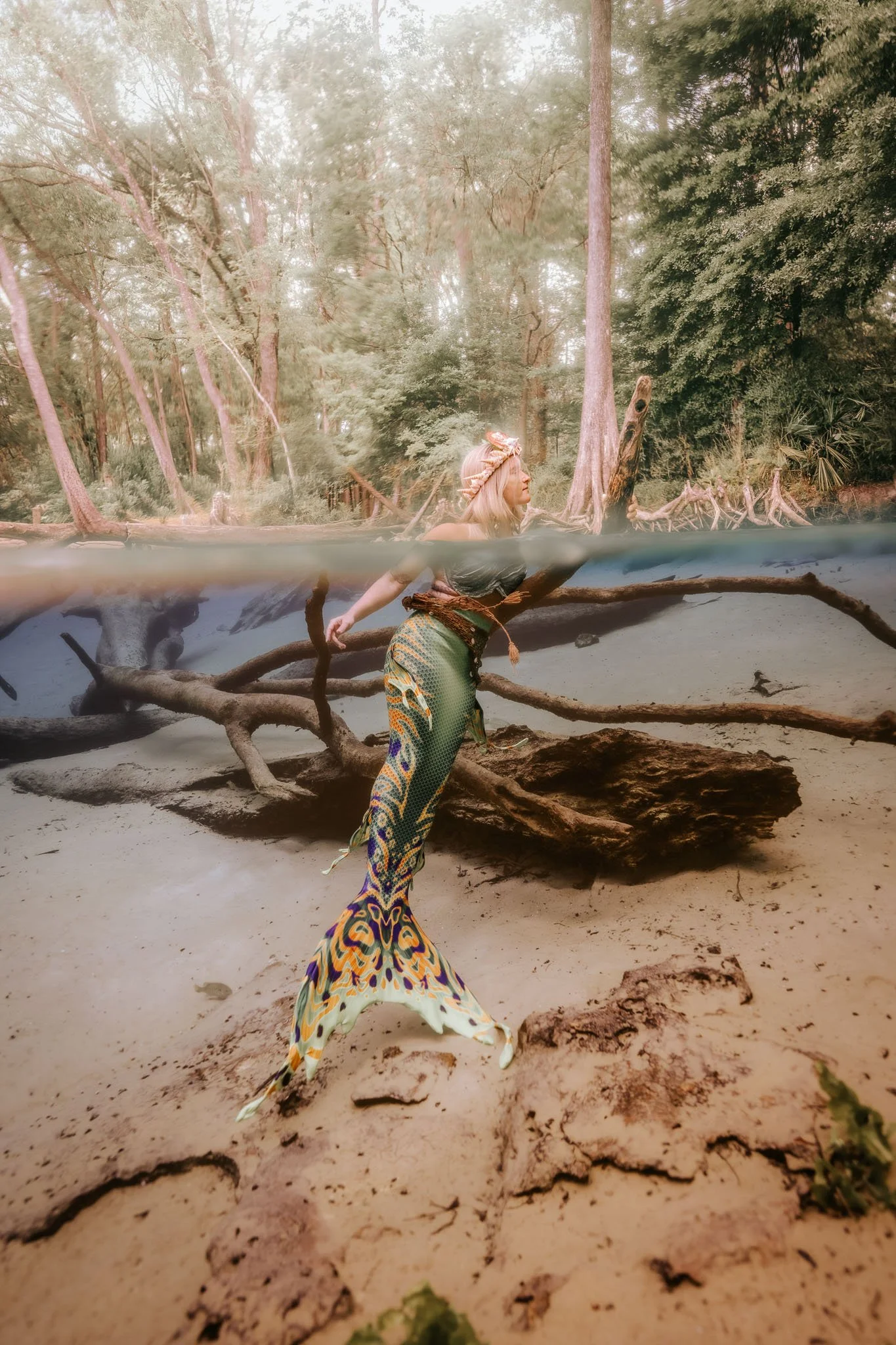 A woman dressed as a mermaid standing on a sandy beach with driftwood and trees in the background, with part of the scene underwater showing her tail and legs.