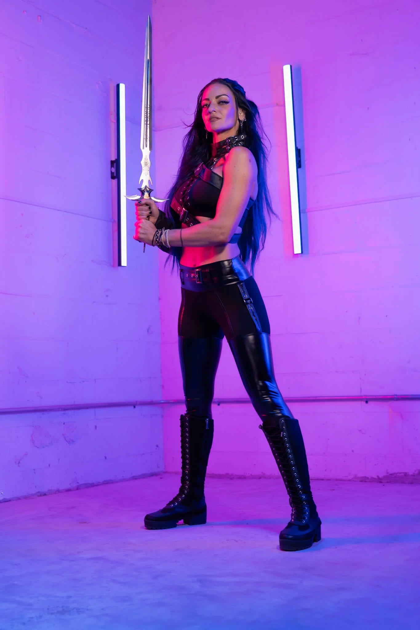 A woman with long dark hair standing in front of a purple-tinted wall holding a sword. She is dressed in black leather, wearing thigh-high lace-up boots and accessories, and has a confident expression.