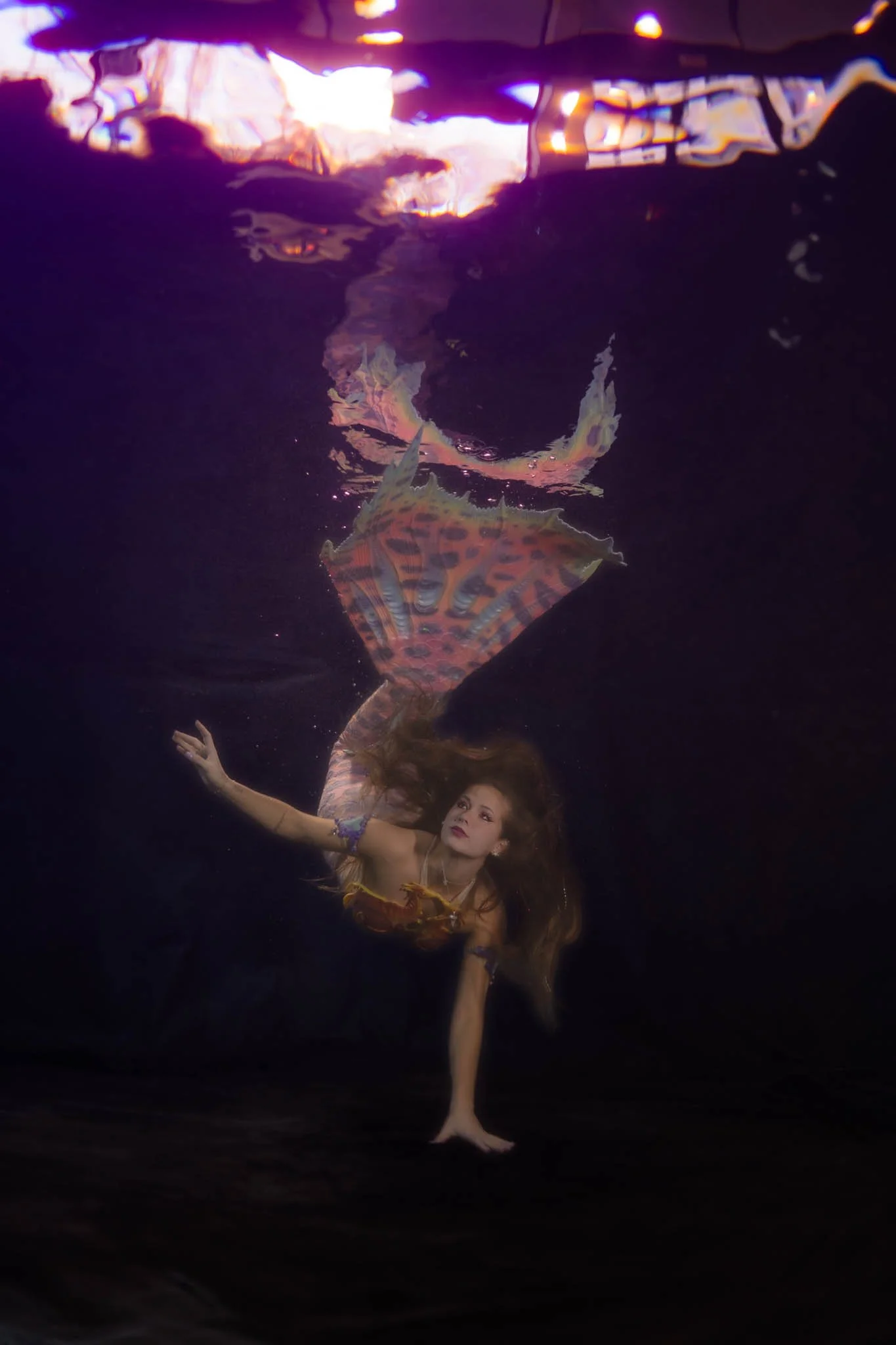 A woman in a colorful outfit is underwater, reaching out with her hand, with a vibrant, patterned fish swimming above her.