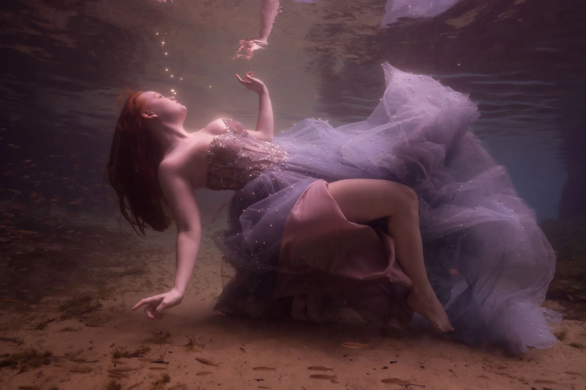 A woman with red hair is underwater, lying on her side, wearing a sparkly pink dress. She is surrounded by water with the surface visible above her. Her eyes are closed, and she appears to be floating gracefully, with her hand raised near her face an