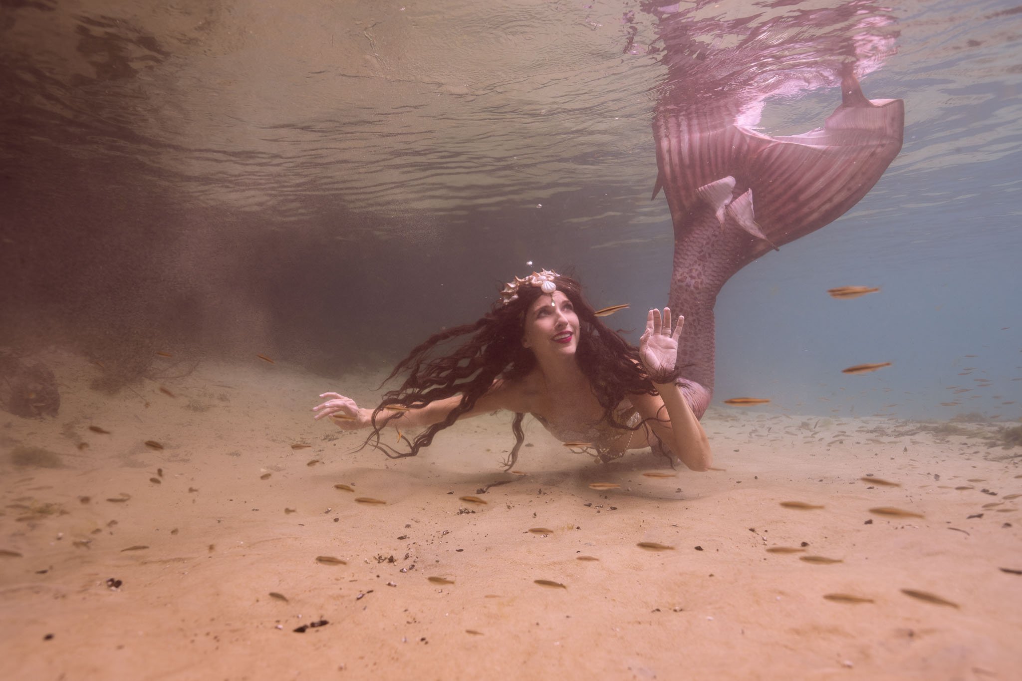 A woman with long dark hair and a crown made of seashells is underwater in a sandy ocean environment, surrounded by small fish, with her right arm raised and waving.
