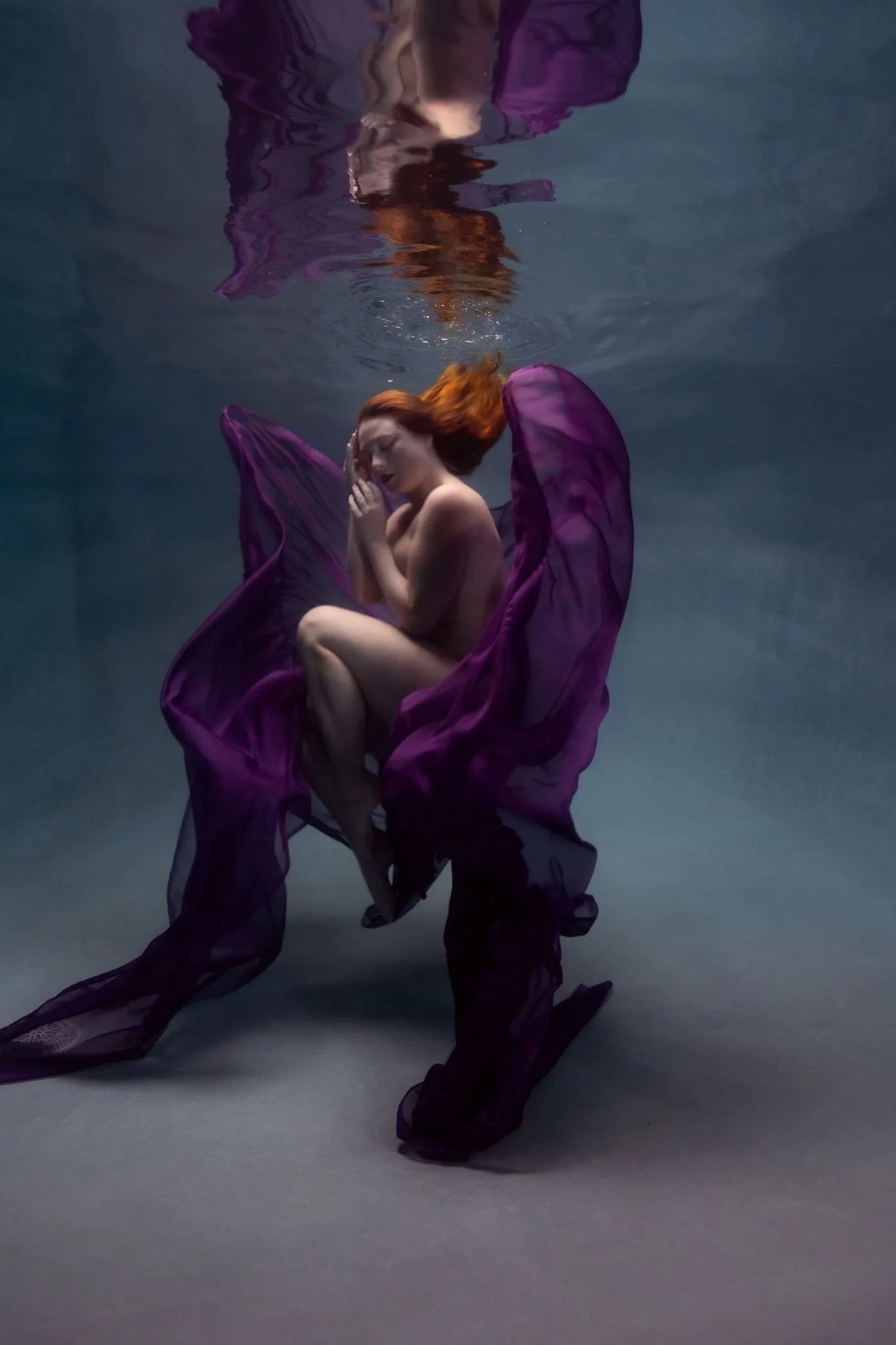 A woman with long red hair, wearing a flowing purple dress, is underwater with her eyes closed and hands gently touching her face, surrounded by soft lighting.