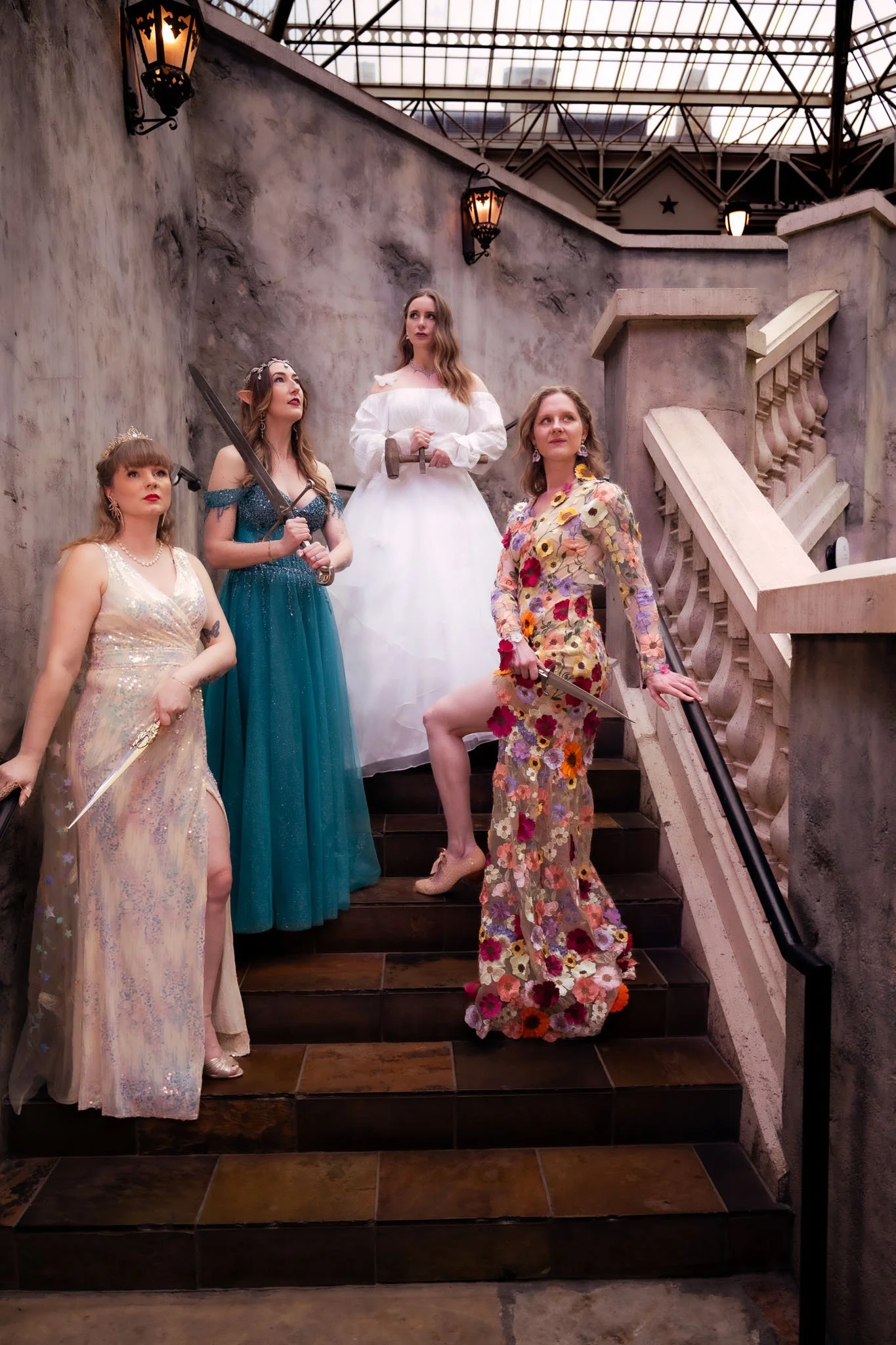 Four women dressed as fantasy characters, including an elf, a princess, a warrior, and a fairy godmother, standing on a staircase inside a building with stone walls and a glass ceiling, holding props such as swords and wearing elaborate costumes and 